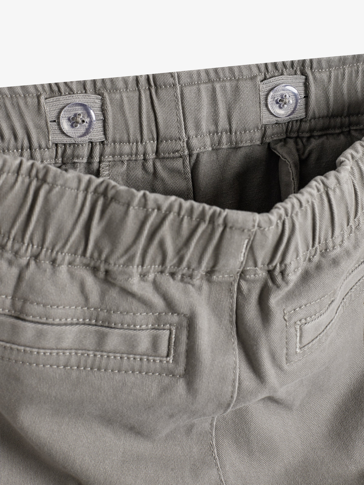 Eine graue Hose zeigt einen elastischen Bund mit Knöpfen zur Größenanpassung. Im Hintergrund sind Gesäßtaschen sichtbar. Die Hose liegt auf einer neutralen Oberfläche.