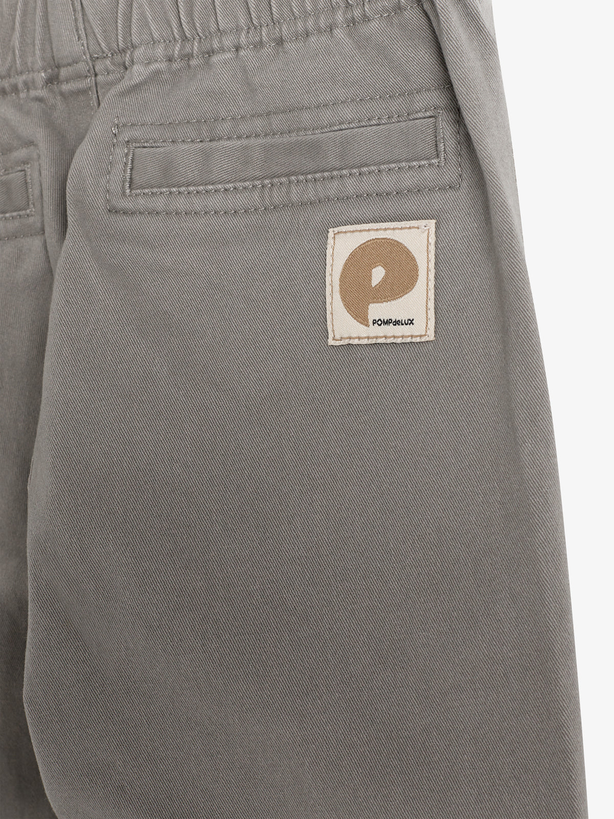 Graue Hose mit Gummibund, rechteckige Gesäßtasche und einem Aufnäher mit einem stilisierten "P" und dem Text "POMPdeLUX" im unteren Bereich. Die Umgebung ist neutral gehalten.