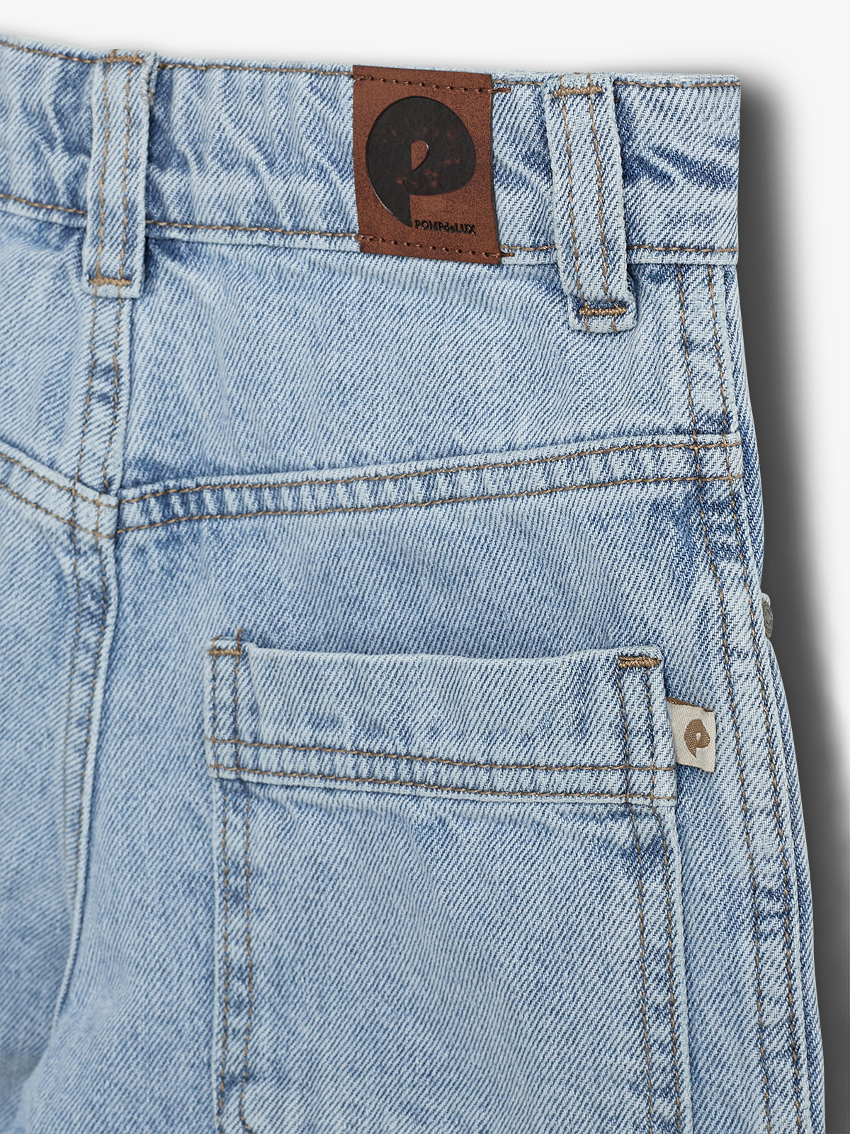 Ein hellblauer Jeansstoff mit Rückentasche zeigt ein braunes Lederschild, auf dem „POMPK&LUX“ steht. Der Stoff ist sorgfältig genäht.