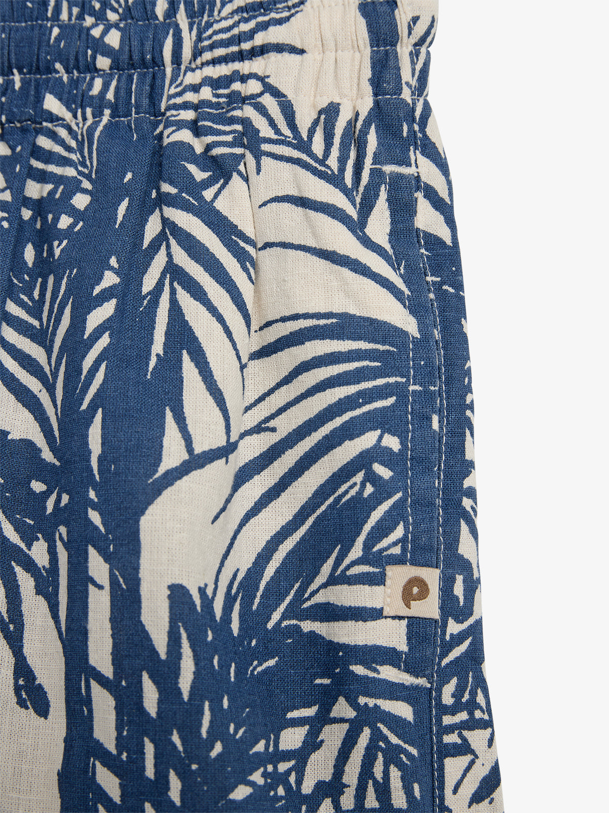 Kurze Hose mit blauem Palmenmuster auf beigem Hintergrund. Elastischer Bund oben, rechts unten ein kleines quadratisches Etikett mit braunem, stilisiertem Buchstaben "p".
