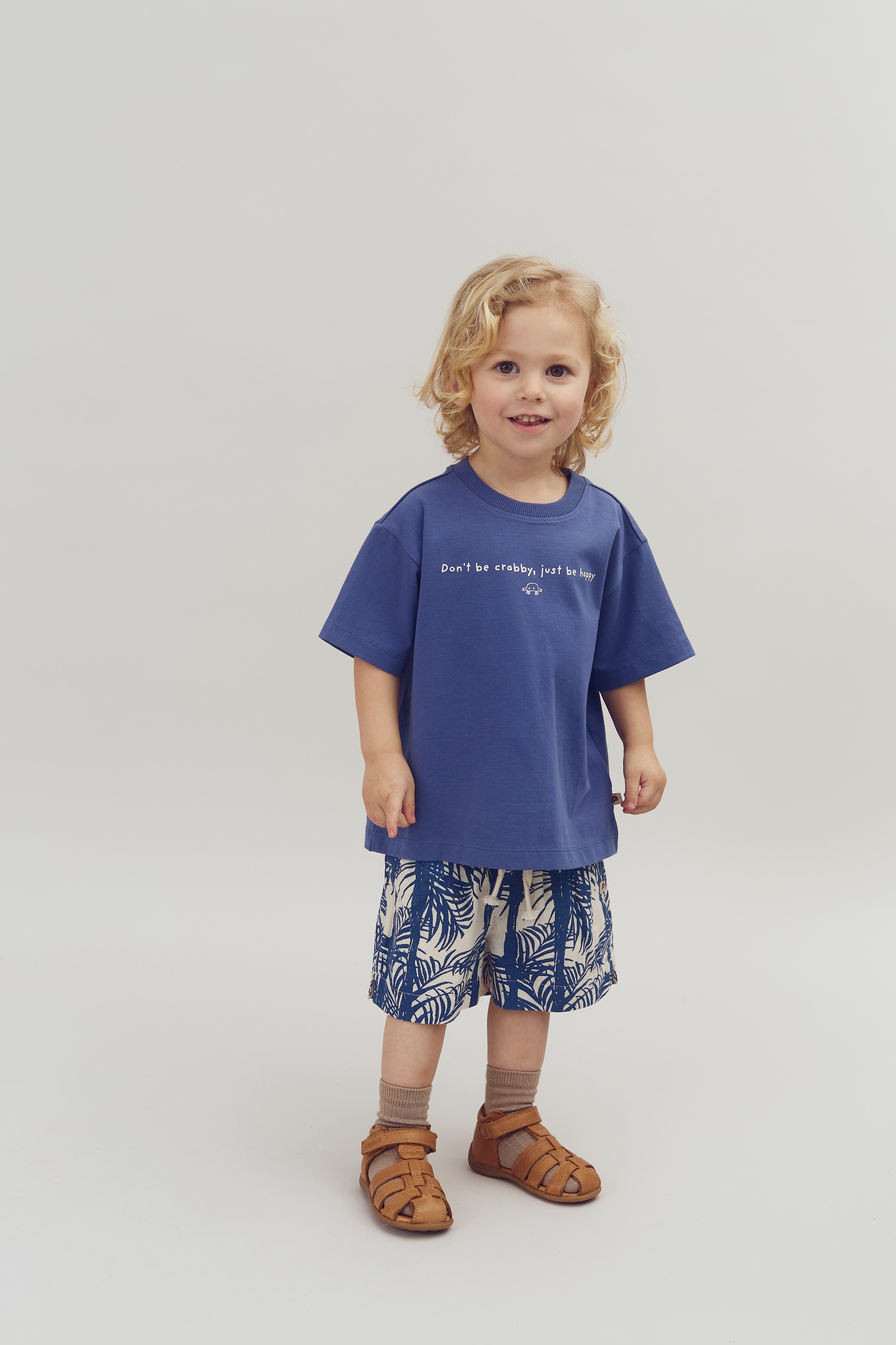 Ein Kind trägt ein blaues T-Shirt mit der Aufschrift "Don't be crabby, just be happy" und kurze Hosen mit Blattmuster. Es steht lächelnd vor einem neutralen Hintergrund.