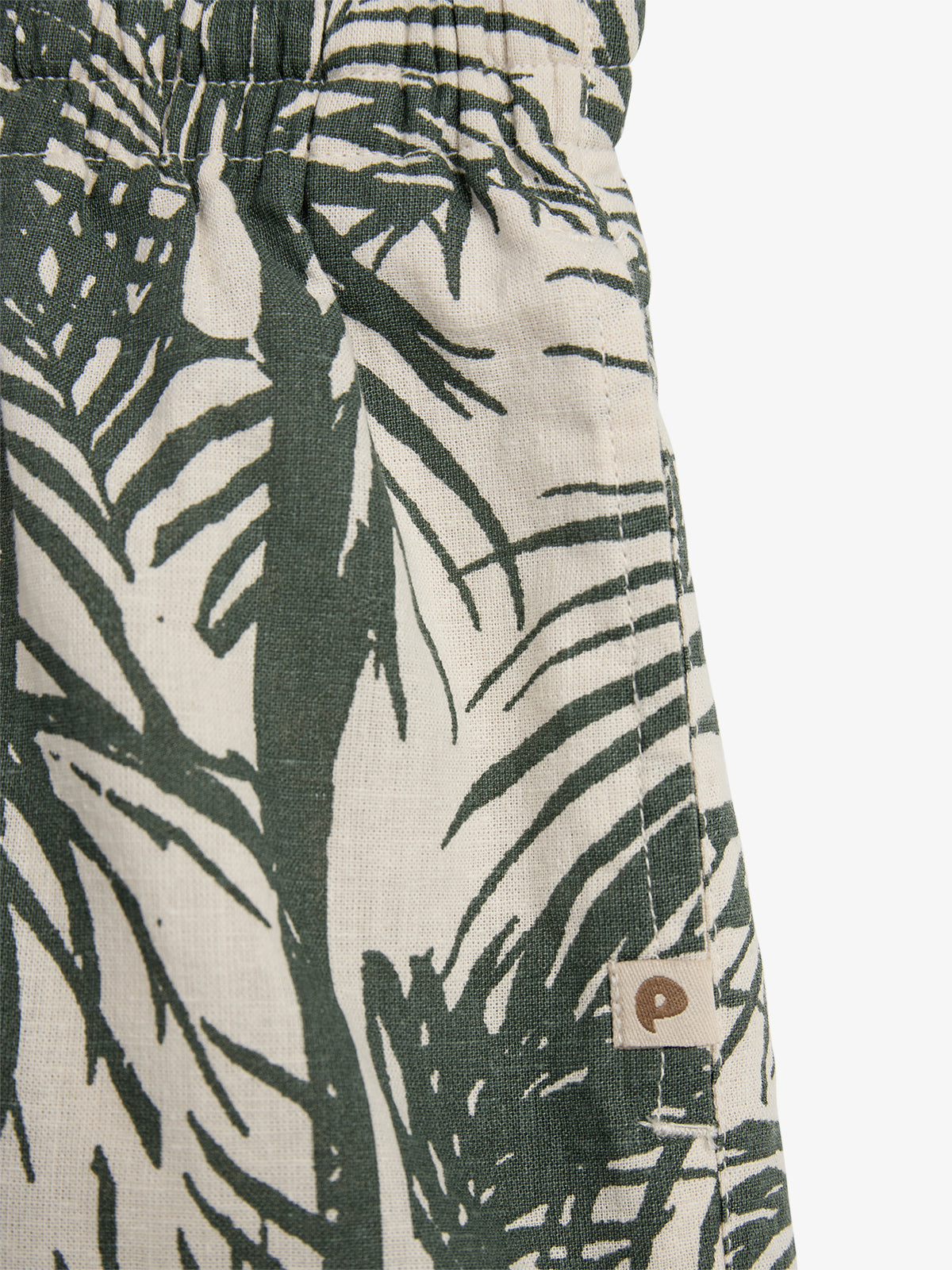 The image shows a close-up of fabric with a green tropical leaf pattern on a beige background. The fabric is gathered at the top, with a sewn tag featuring a small, brown 'P'.