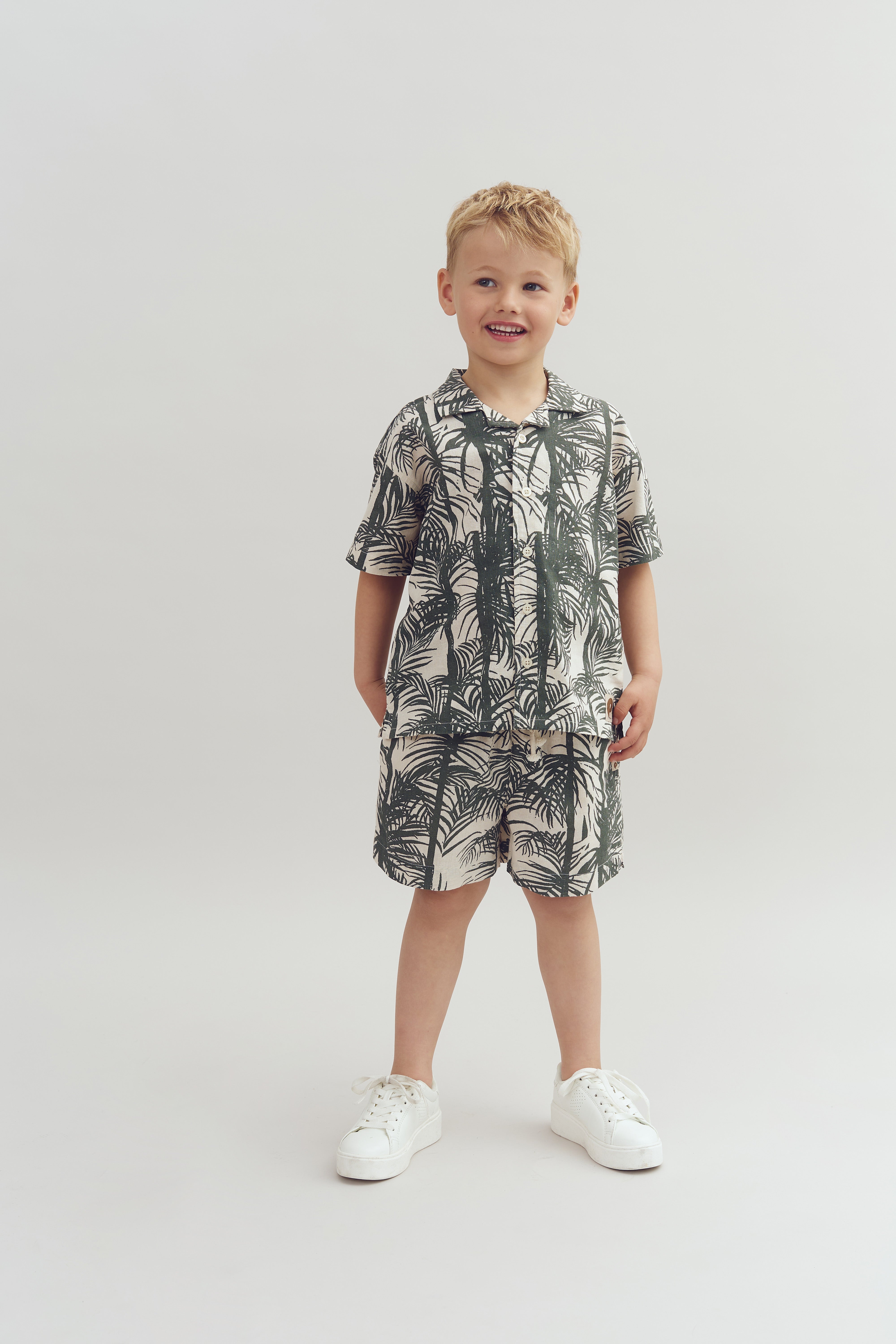 Child stands smiling, wearing a matching tropical leaf-patterned shirt and shorts set with white sneakers. The background is a plain, light color.