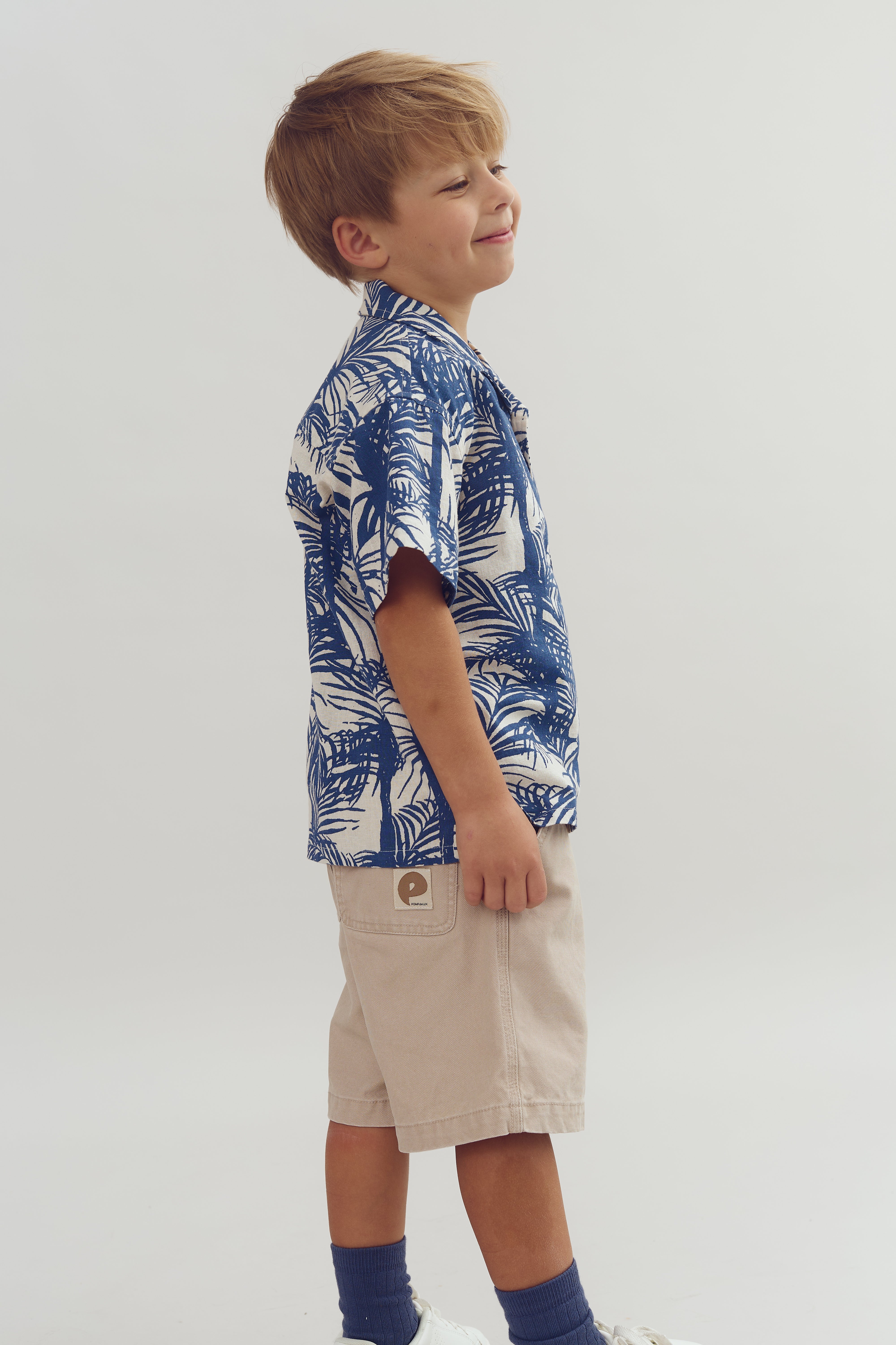 Junge mit blau-weißem, tropisch gemustertem Hemd lächelt zur Seite. Er trägt beige Shorts mit aufgesetztem Etikett und blaue Socken. Der Hintergrund ist neutral.