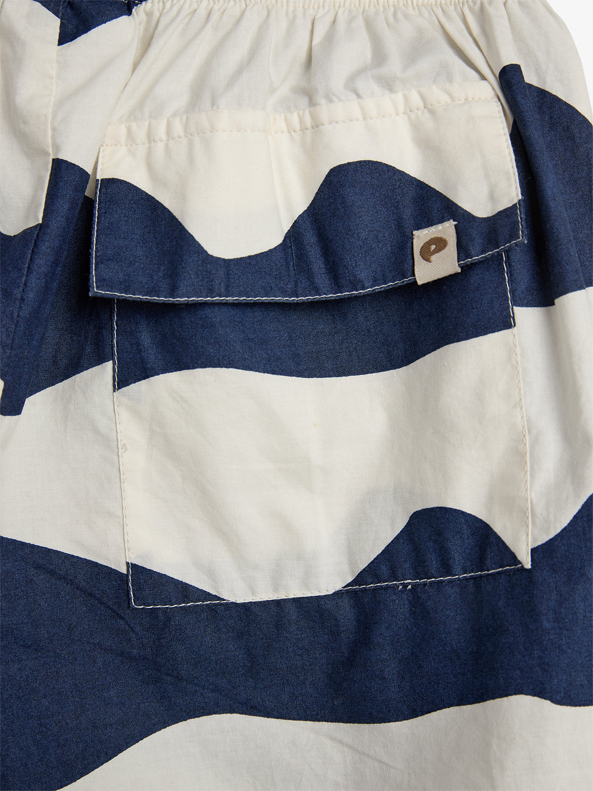 Tasche mit dunkelblau-weißen Streifenmuster, an einem Kleidungsstück angenäht; beige Etikett mit einem kleinen, dunkelbraunen Logo in der oberen rechten Ecke; umgeben von ähnlichem Streifenmuster.