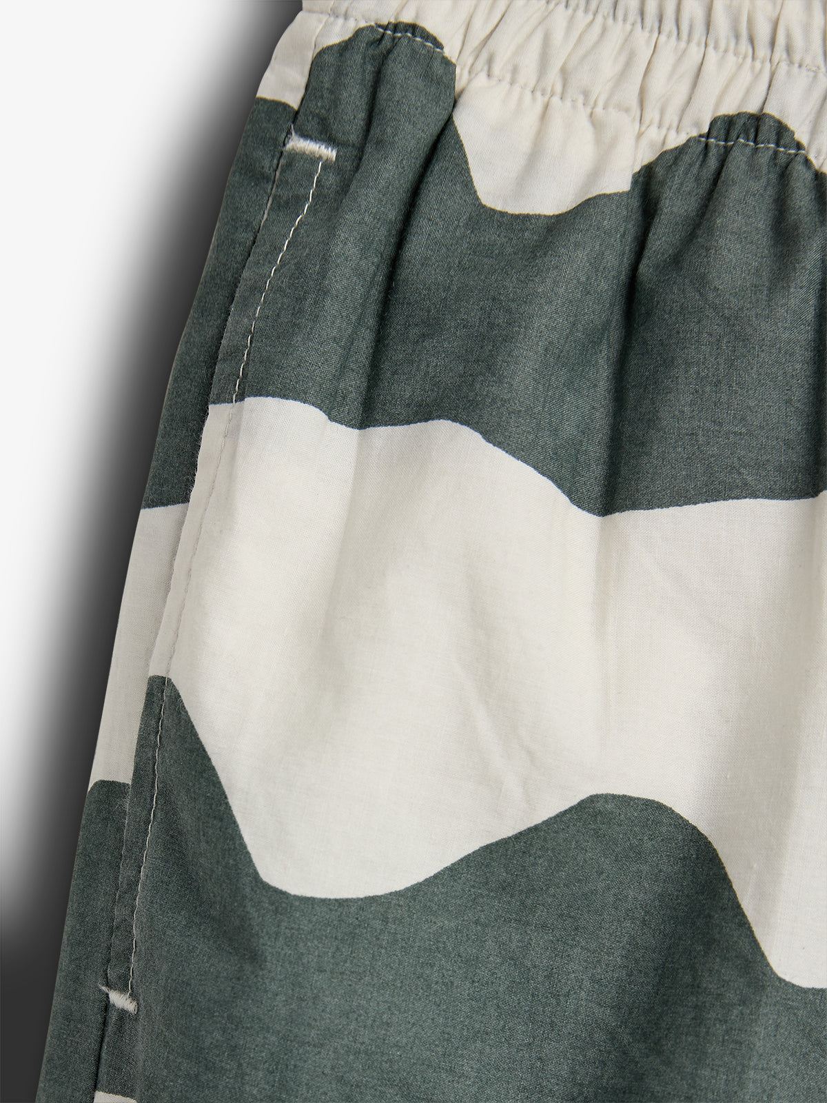 Eine gestreifte Hose zeigt grün-weiße Wellenmuster. Die elastische Taille ist sichtbar, während die Nähte entlang der Kante verlaufen. Der Hintergrund ist einfärbig grau.