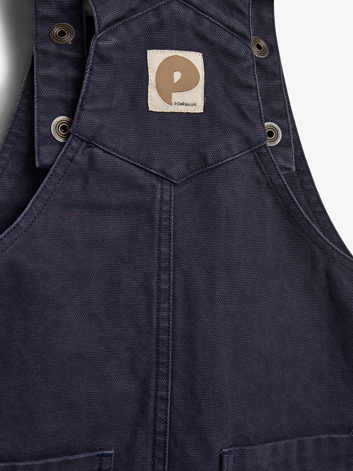 Dark blue bib overalls feature metal grommets and a tan patch with "POMPdeLUX" and an abstract "P" logo, set against a plain background.