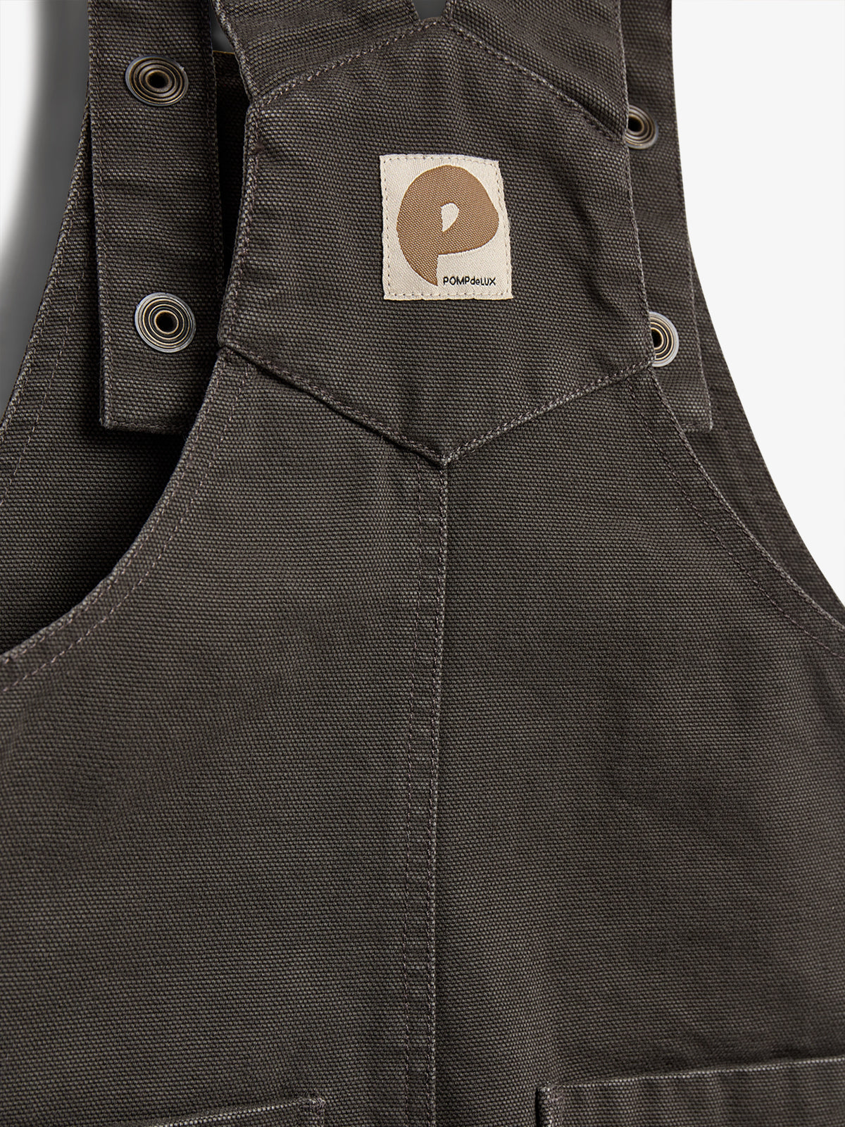 Dark fabric garment featuring a brown logo patch with a stylized "P" and text "POMPdeLUX." Metal eyelets appear on the straps, indicating functionality and design elements.