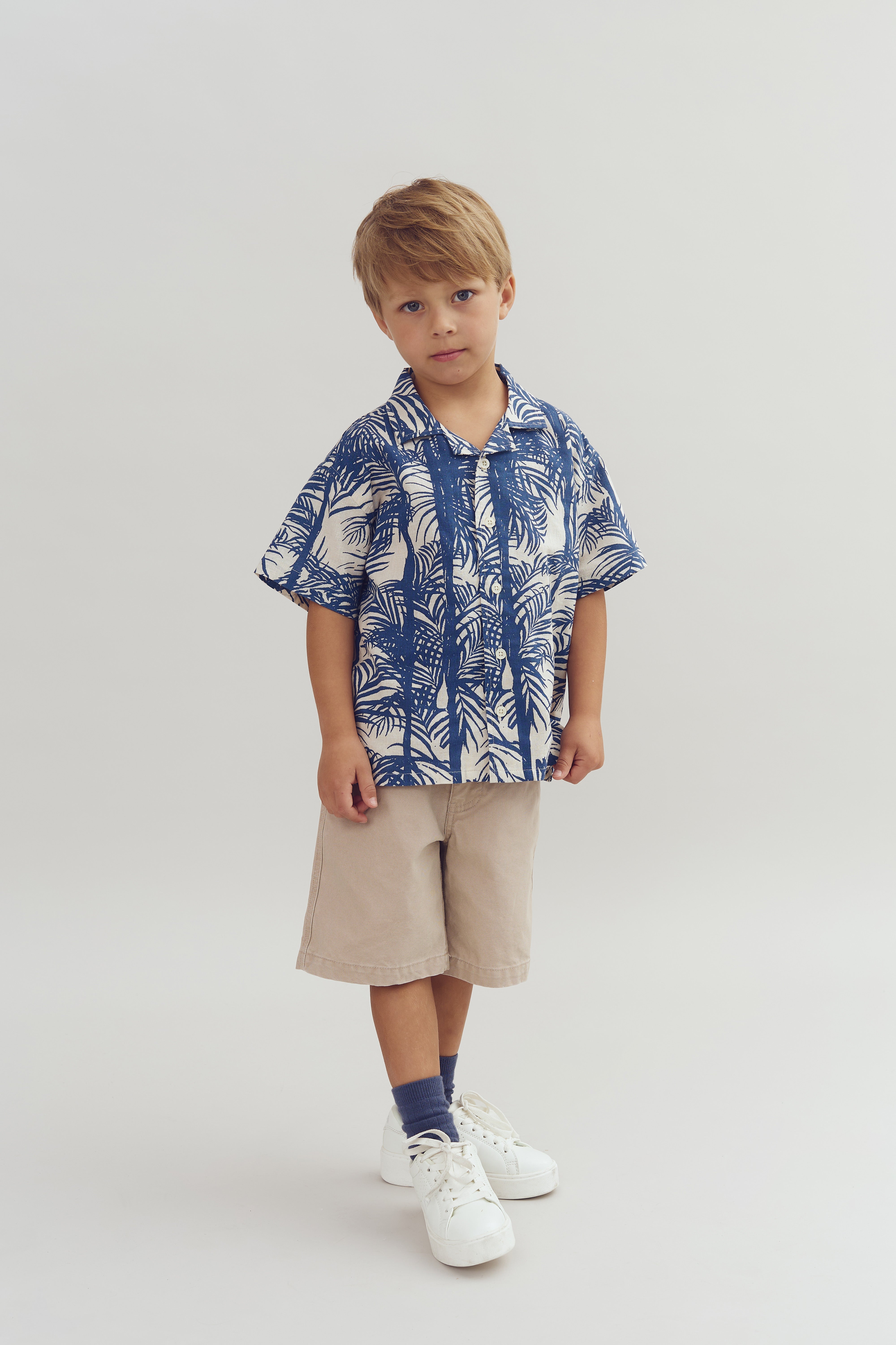 Ein Junge steht ruhig, trägt ein blau-weißes Hemd mit Blattmuster, beige Shorts und weiße Turnschuhe. Der Hintergrund ist einfarbig hell.
