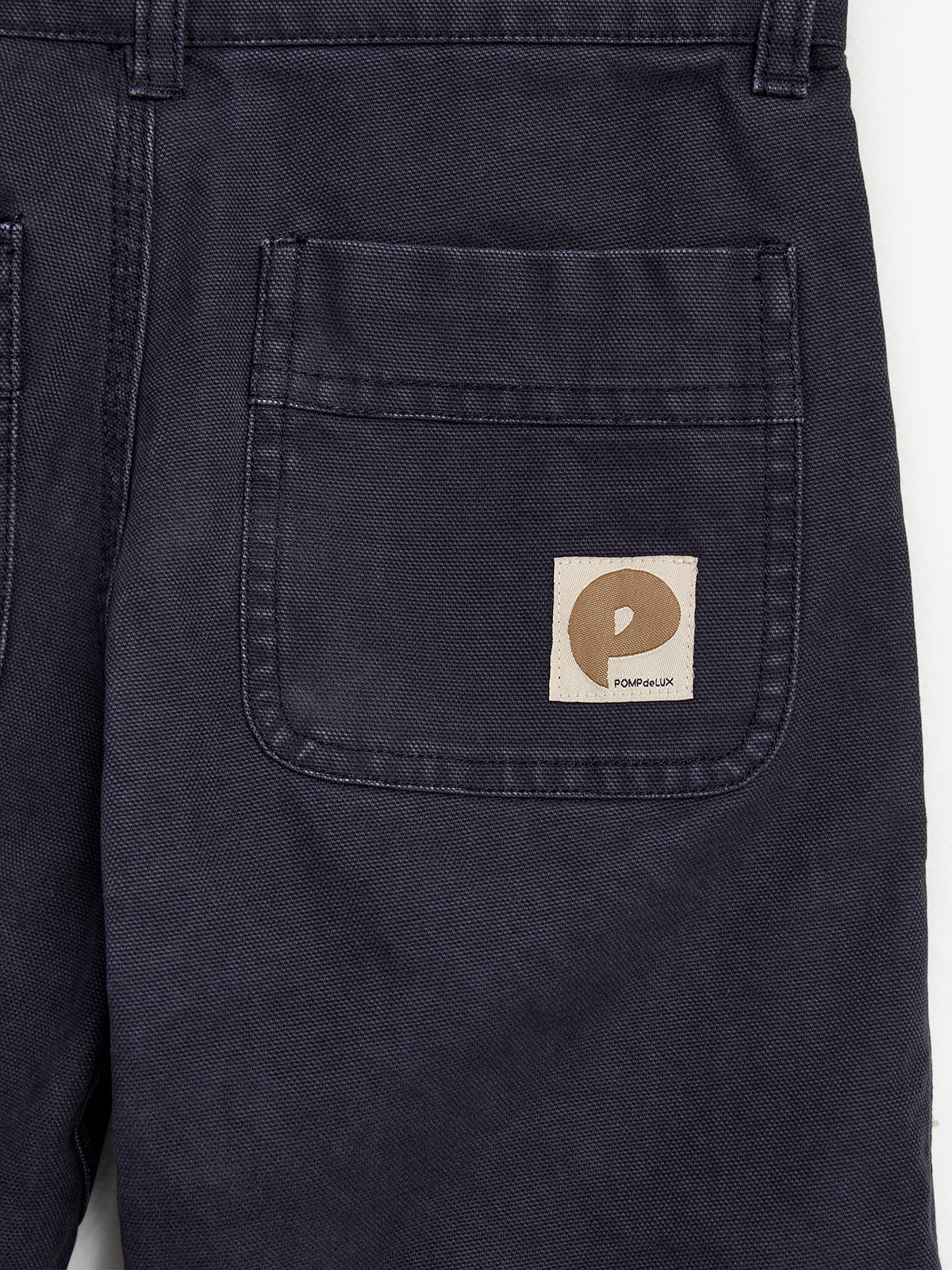 Dunkelblaue Hose mit einer Tasche und einem Etikett. Das Etikett zeigt ein stilisiertes "P" und den Text "POMPdeLUX". Baumwollstoff umgibt die Szene.