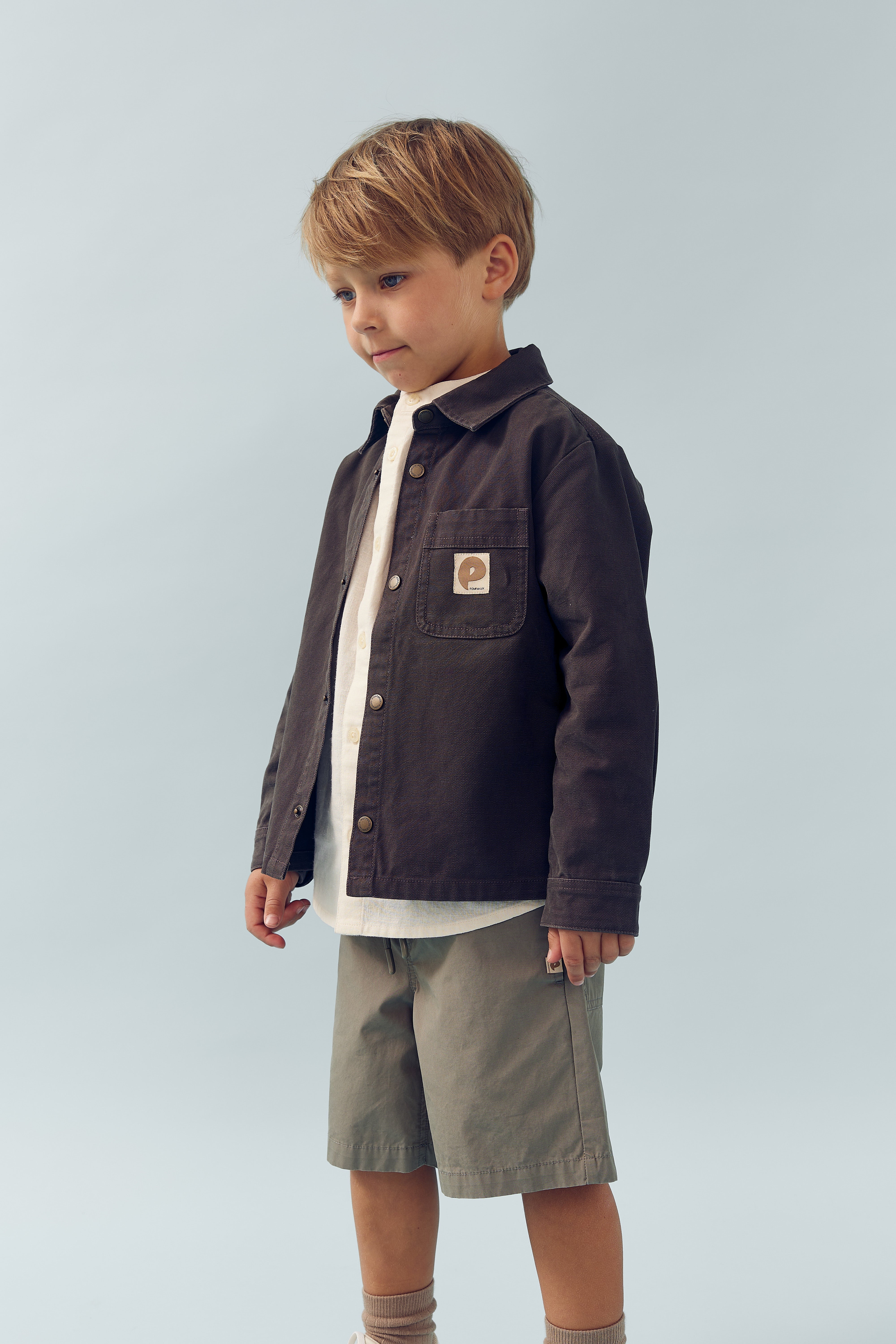 Ein Kind steht ruhig in einem hellen Raum. Es trägt eine dunkle Jacke über einem hellen Hemd und olivfarbene Shorts. Ein Emblem mit "e" ist auf der Jackentasche.