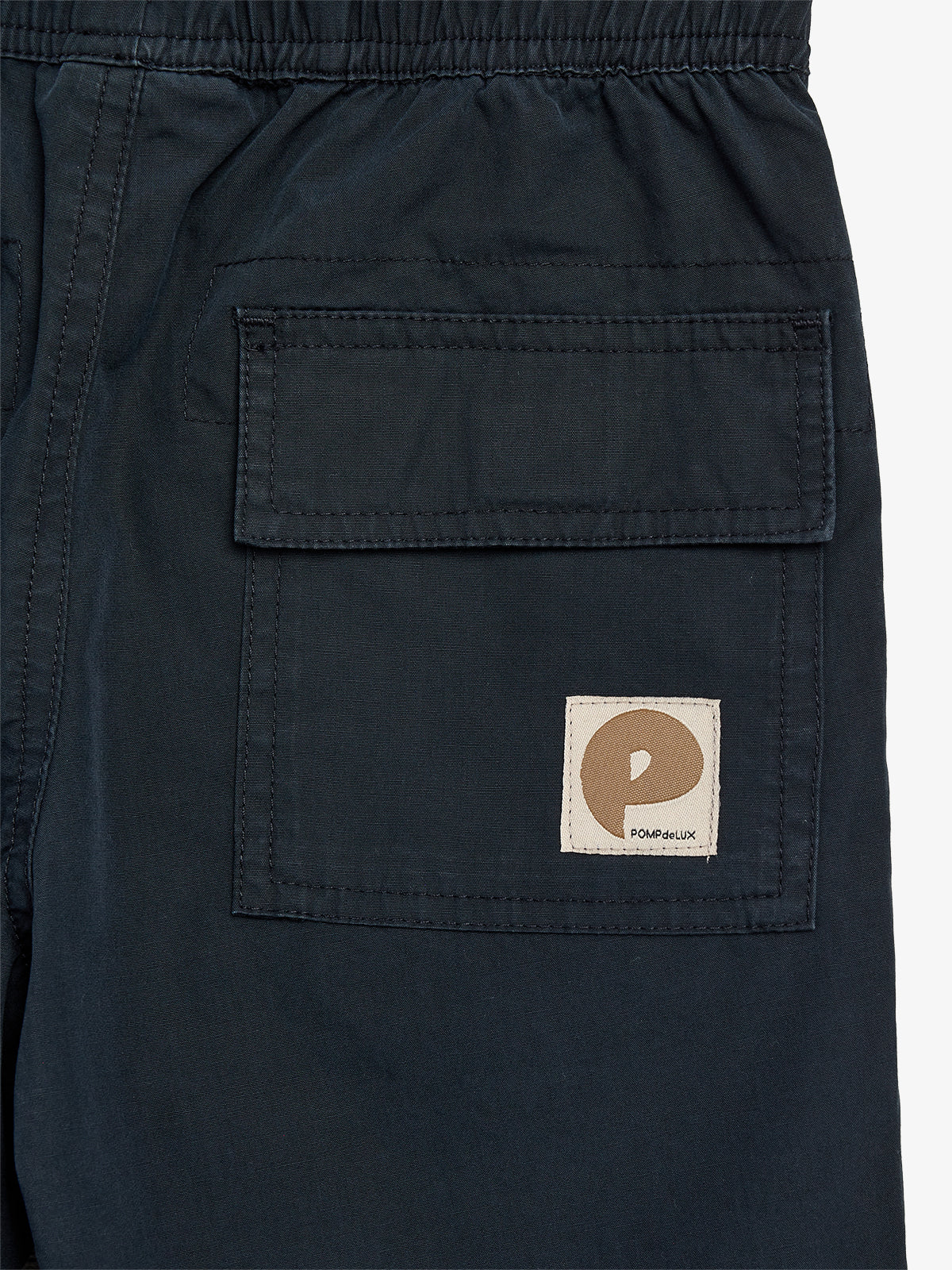 Tasche auf einer dunkelblauen Shorts mit einer Patte und einem aufgenähten Etikett. Etikett zeigt ein braunes Logo und den Text "POMPdeLUX". Umgebung ist schlicht und neutral.