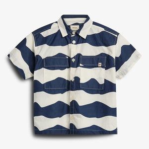 Ein kurzärmeliges Hemd mit wellenförmigen, horizontalen blauen und weißen Streifen. Zwei Brusttaschen mit Knöpfen ergänzen das Design. Die Umgebung ist neutral.