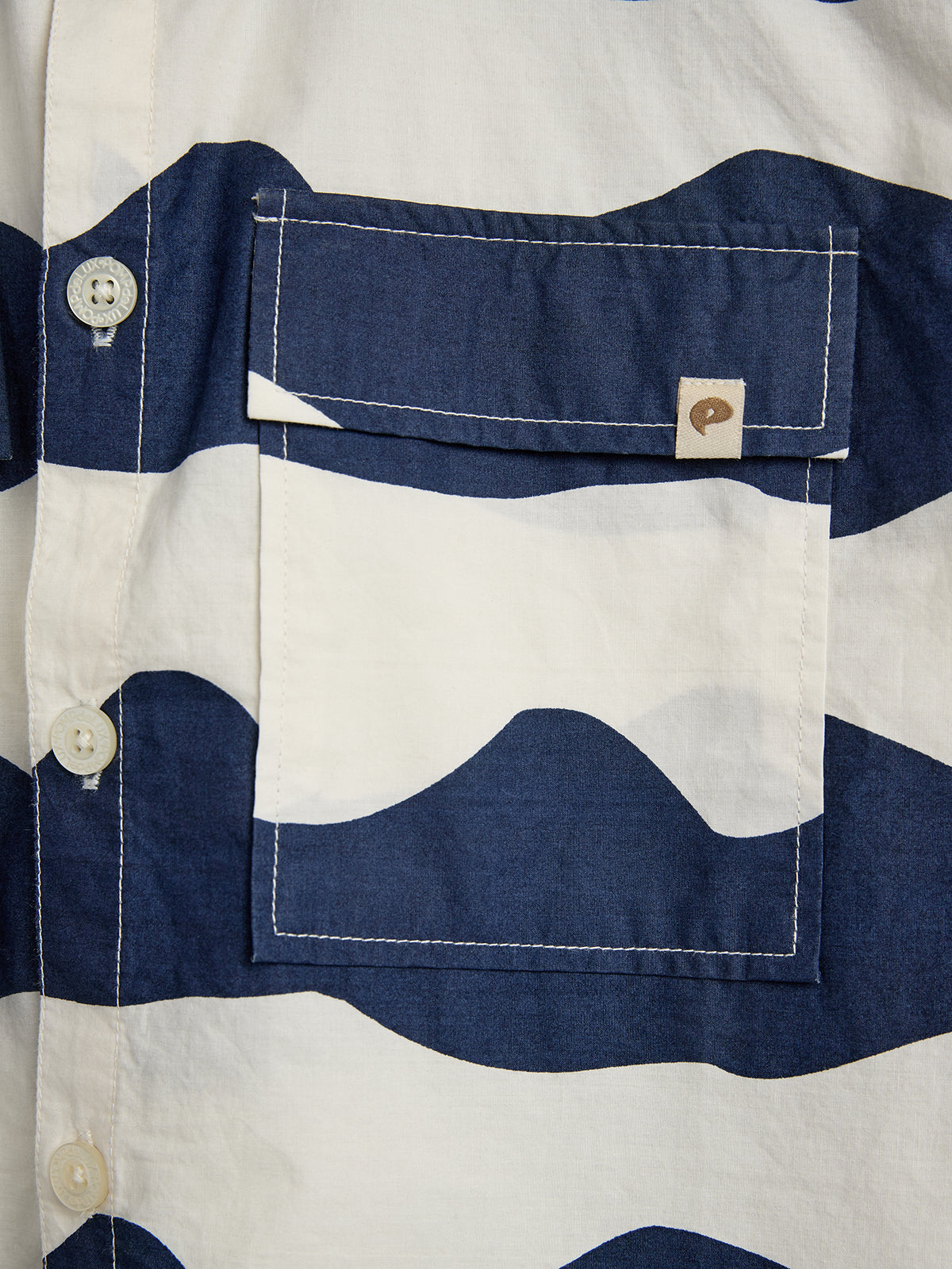 Eine blau-weiß gestreifte Hemdtasche mit Knopfverschluss und Wellenmuster auf beigefarbenem Stoff. Daneben sind drei weiße Knöpfe zu sehen. Ein kleines Label ist an der Tasche befestigt.