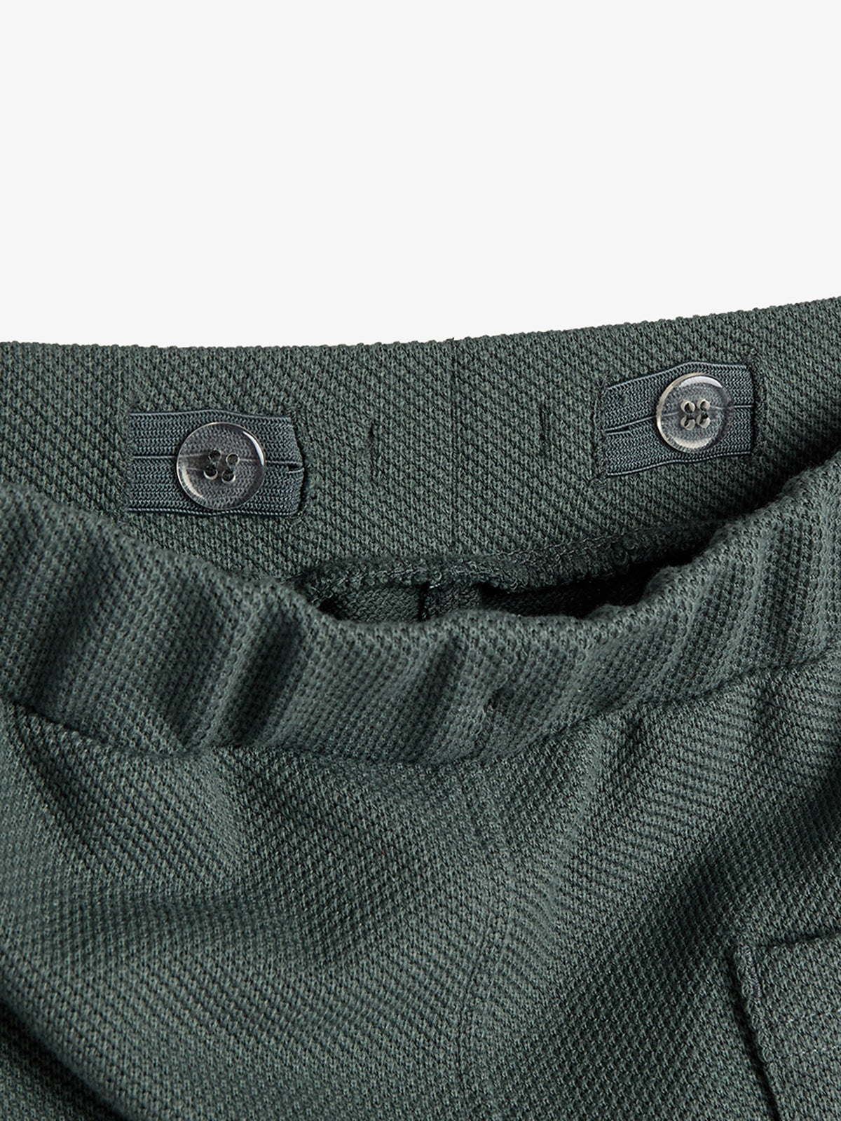 The garment's waistband, made of textured green fabric, features an adjustable elastic band with two attached buttons. The context is a close-up view, focusing on waist detail.