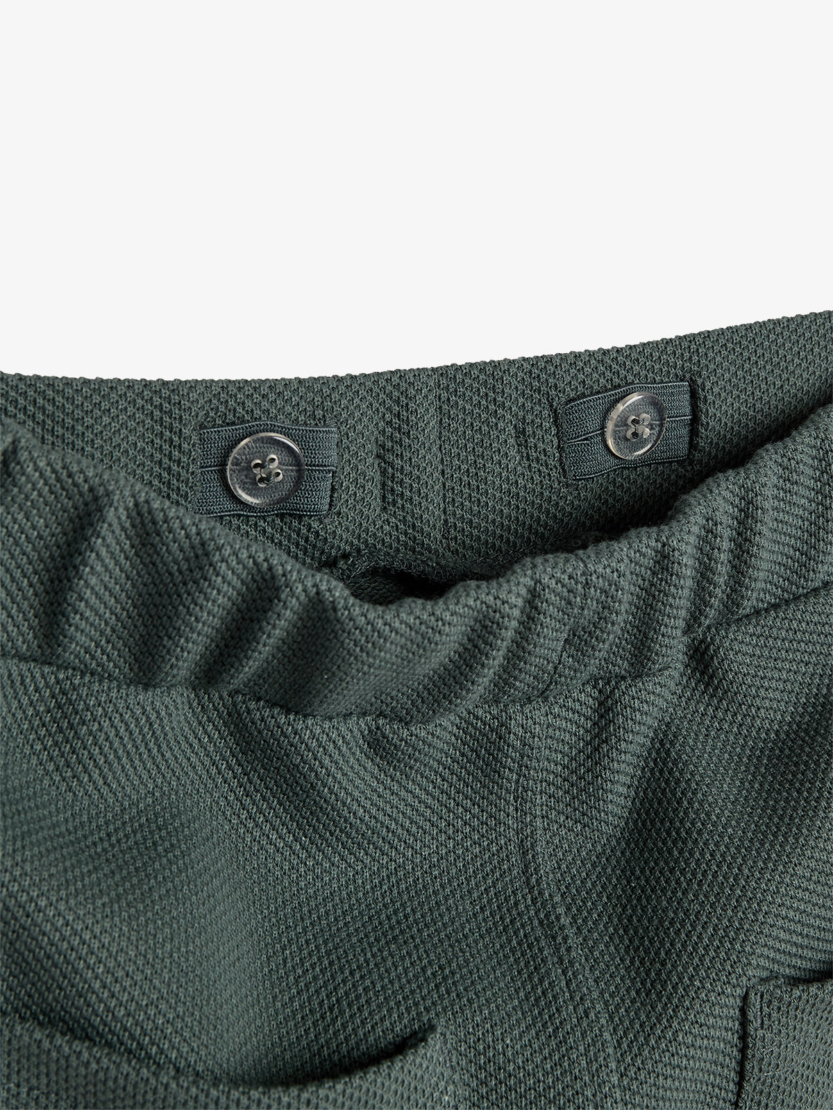 Dunkelgrüne Hose zeigt eine elastische Taille mit zwei schwarzen Knöpfen. Der Stoff hat eine strukturelle, grobmaschige Oberfläche. Im Bildhintergrund ist ein weißer, leerer Raum zu sehen.