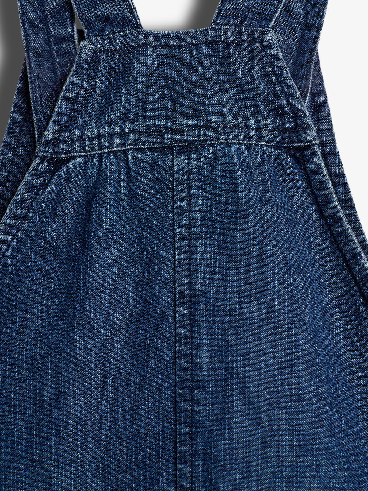 Dunkelblauer Jeansstoff zeigt die Vorderseite eines Latzkleides. Der Stoff ist durchgehend genäht, mit zwei Trägern oben, in einer Studioaufnahme ohne sichtbaren Hintergrund.