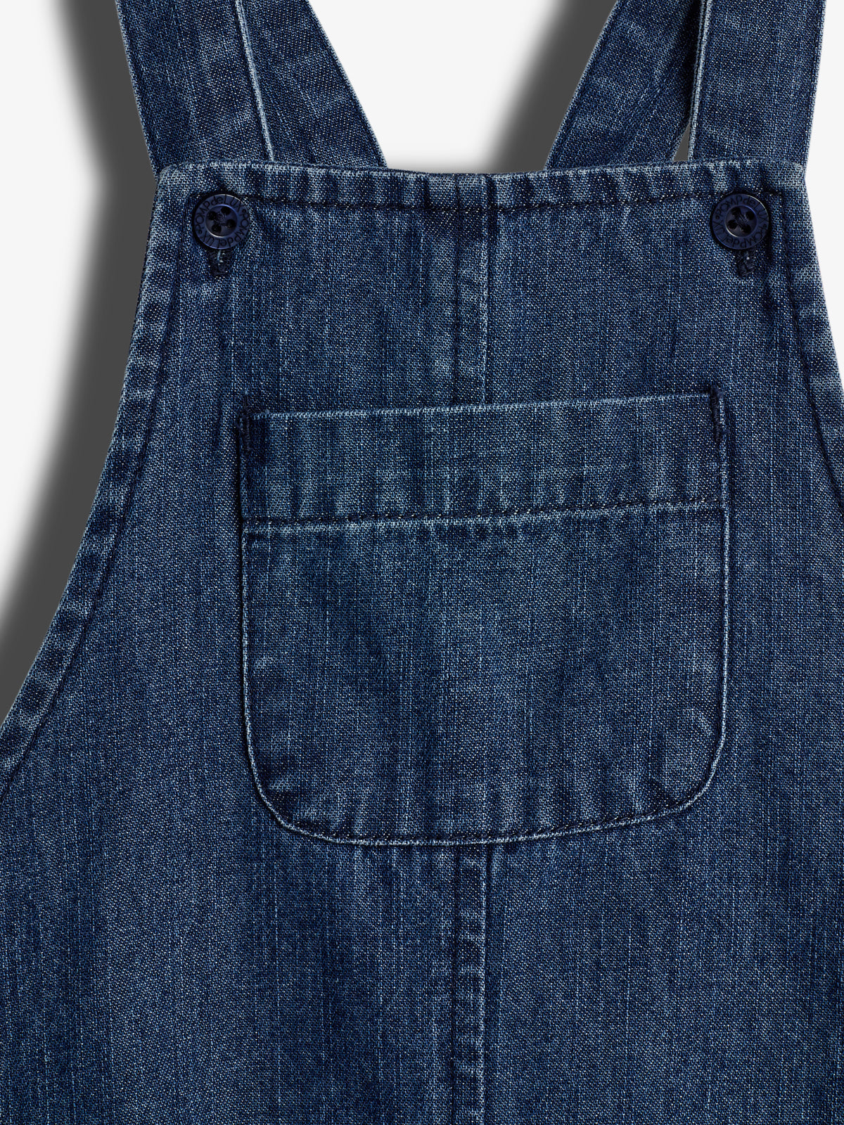 Ein dunkelblaues Jeanslätzchen mit Knöpfen oben, Taschen vorne, sitzt im starken Licht mit Schatten an den Rändern, wirkt strukturiert und robust.