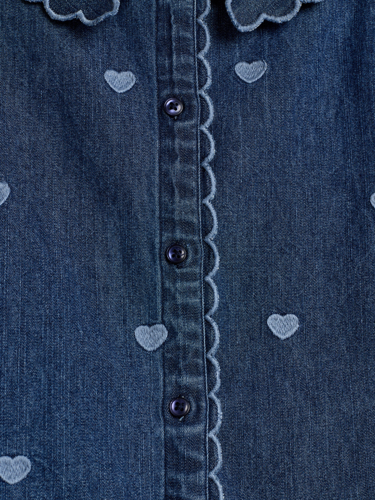 Ein blaues Jeanshemd mit Herzmustern und einer Knopfleiste aus schwarzen Knöpfen. Die Naht entlang der Knopfleiste ist geschwungen. Die Herzen sind gleichmäßig verteilt.