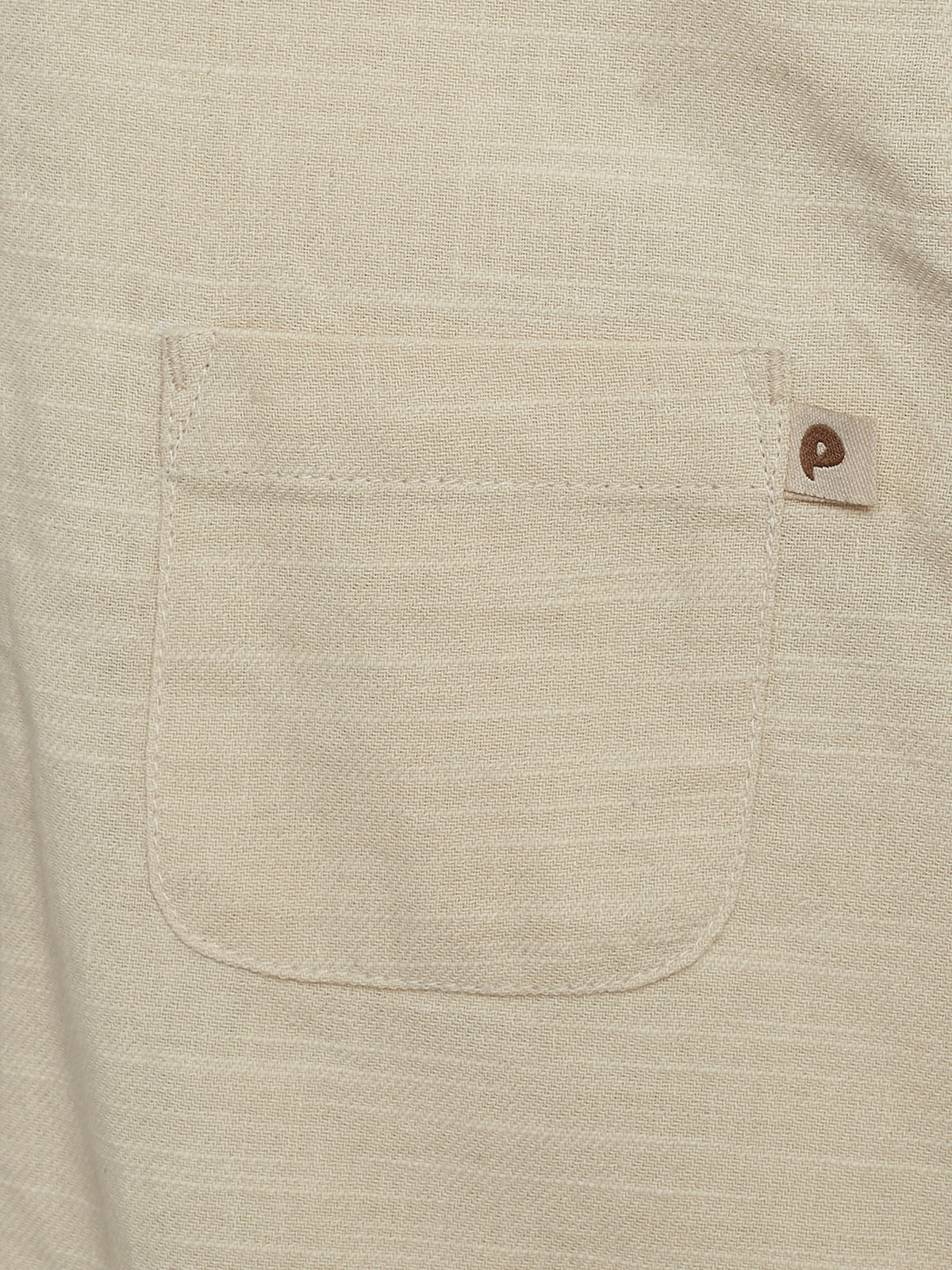 Eine beigefarbene Tasche ist auf einem Stoff sichtbar, links unten genäht. Rechts oben befindet sich ein kleines Etikett mit einem braunen Symbol darauf, eingebettet in ein strukturiertes Gewebe.