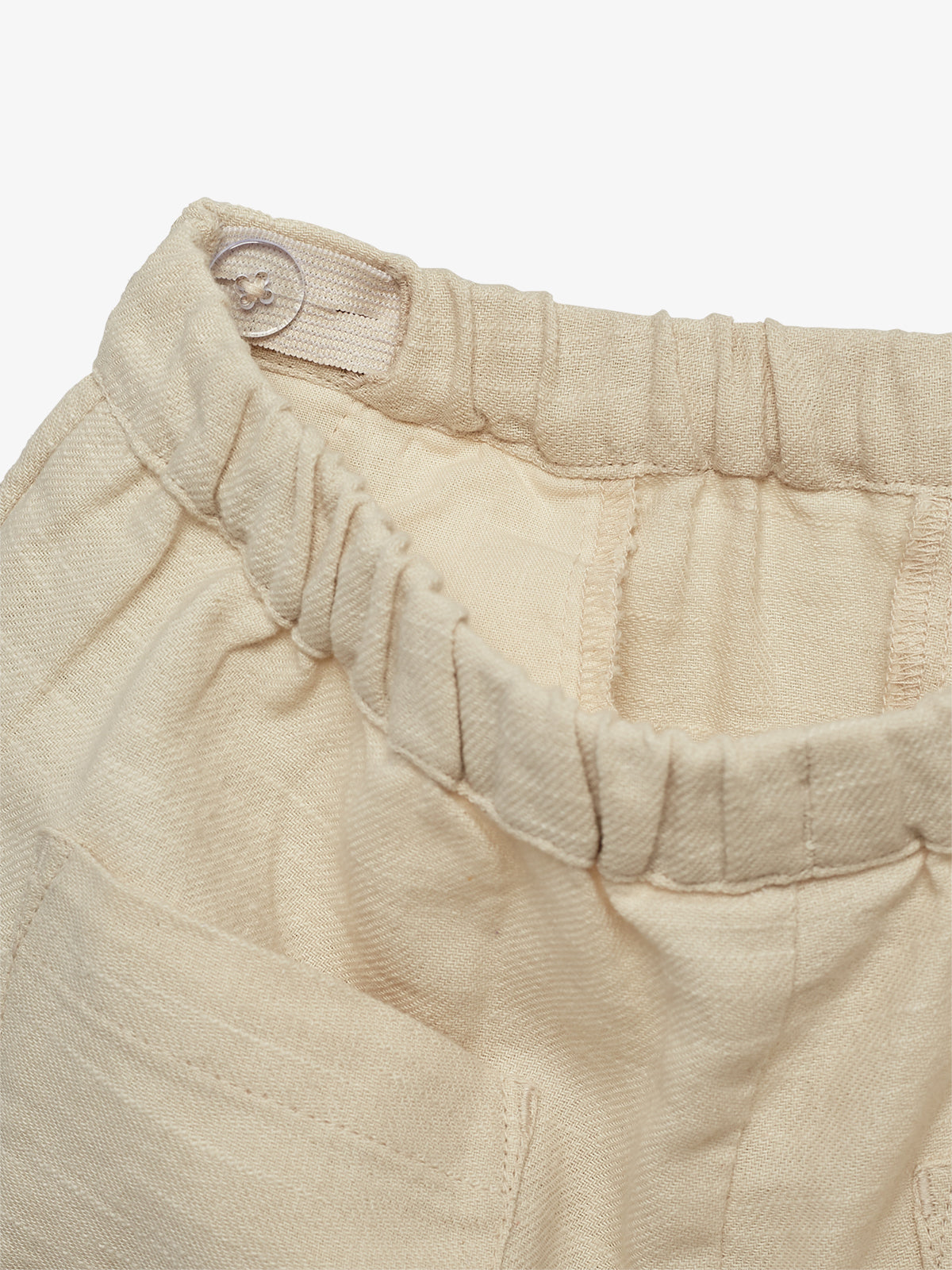 Beige Stoffhose mit elastischem Bund, teilweise sichtbar in Nahaufnahme. Ein kleiner Knopf ist am Bund befestigt. Die Umgebung ist einfarbig und schlicht gehalten.