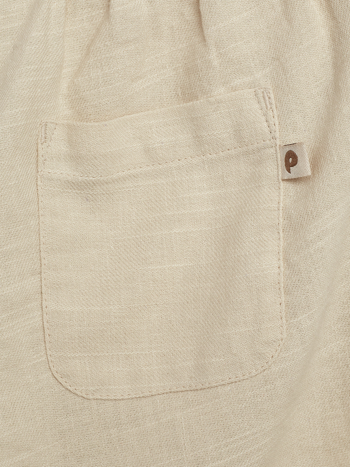Eine beige Textiltasche ist auf einem Stoffstück genäht. Eine kleine Lasche mit einem braunen, stilisierten „p“ ist am Rand der Tasche befestigt.