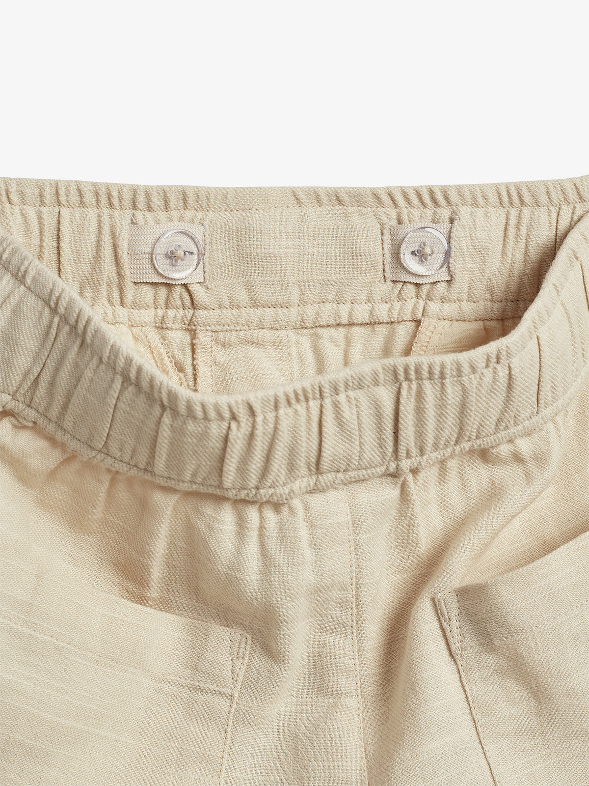Hose mit elastischer Taille und zwei sichtbaren Knöpfen zur Anpassung. Der Stoff ist beige und strukturiert, mit angedeuteten Taschen daneben, auf einem neutralen Hintergrund.