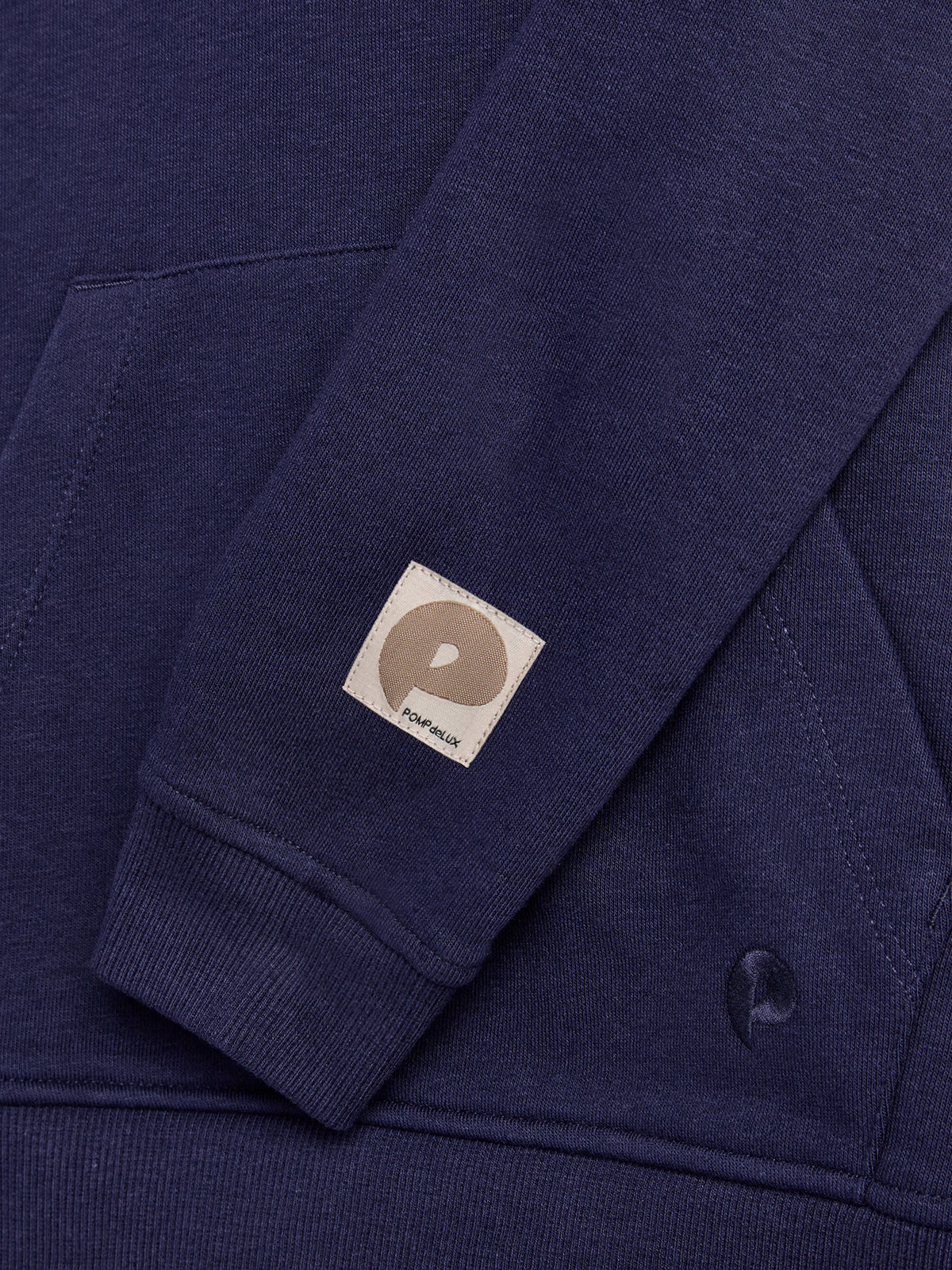 A dark blue sweatshirt features a folded sleeve with a small beige patch labeled "POMPdeLUX." The sleeve rests across the front pocket area, set against the same fabric.