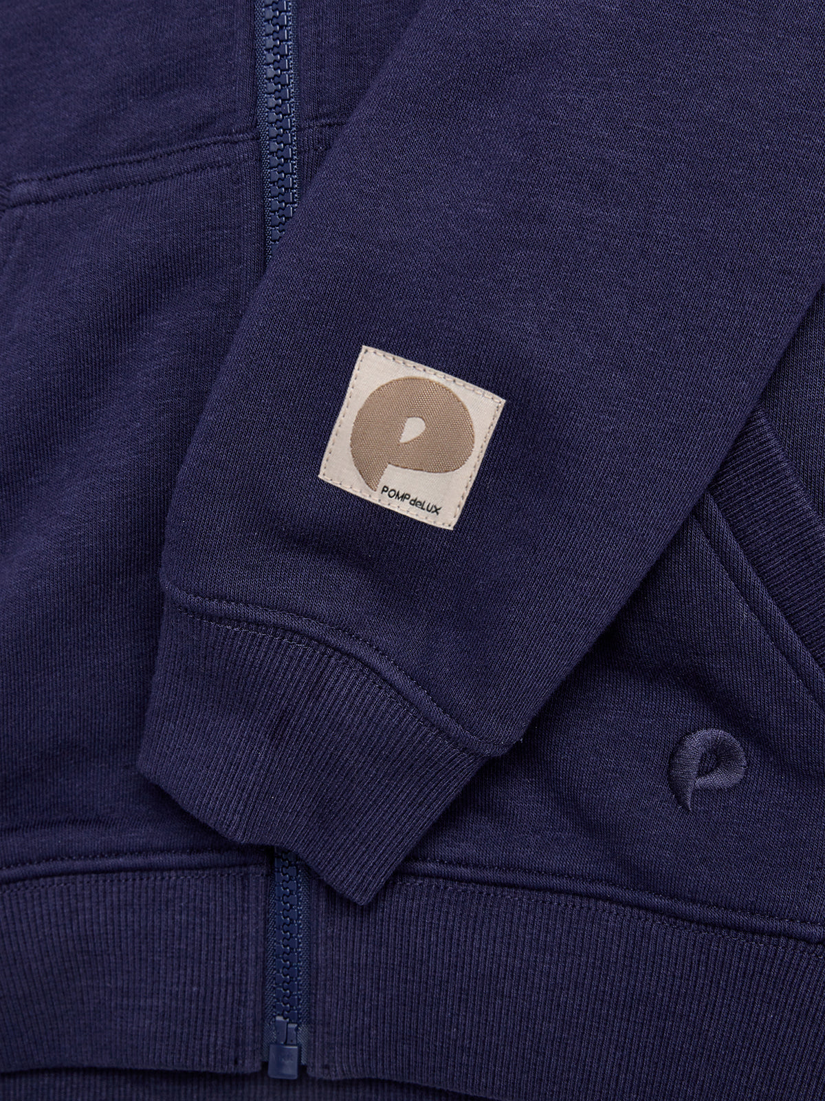 A dark blue zip-up hoodie features a beige patch with a stylized "P" and the text "POMP de LUX," set against a matching ribbed cuff and background.