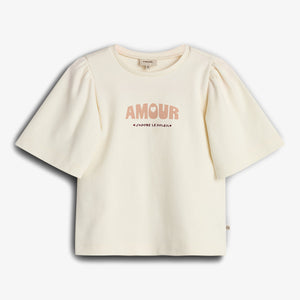 Ein cremefarbenes T-Shirt zeigt in der Mitte den Text "AMOUR J'ADORE LE SOLEIL". Es hat kurze, leicht puffige Ärmel und befindet sich vor weißem Hintergrund.