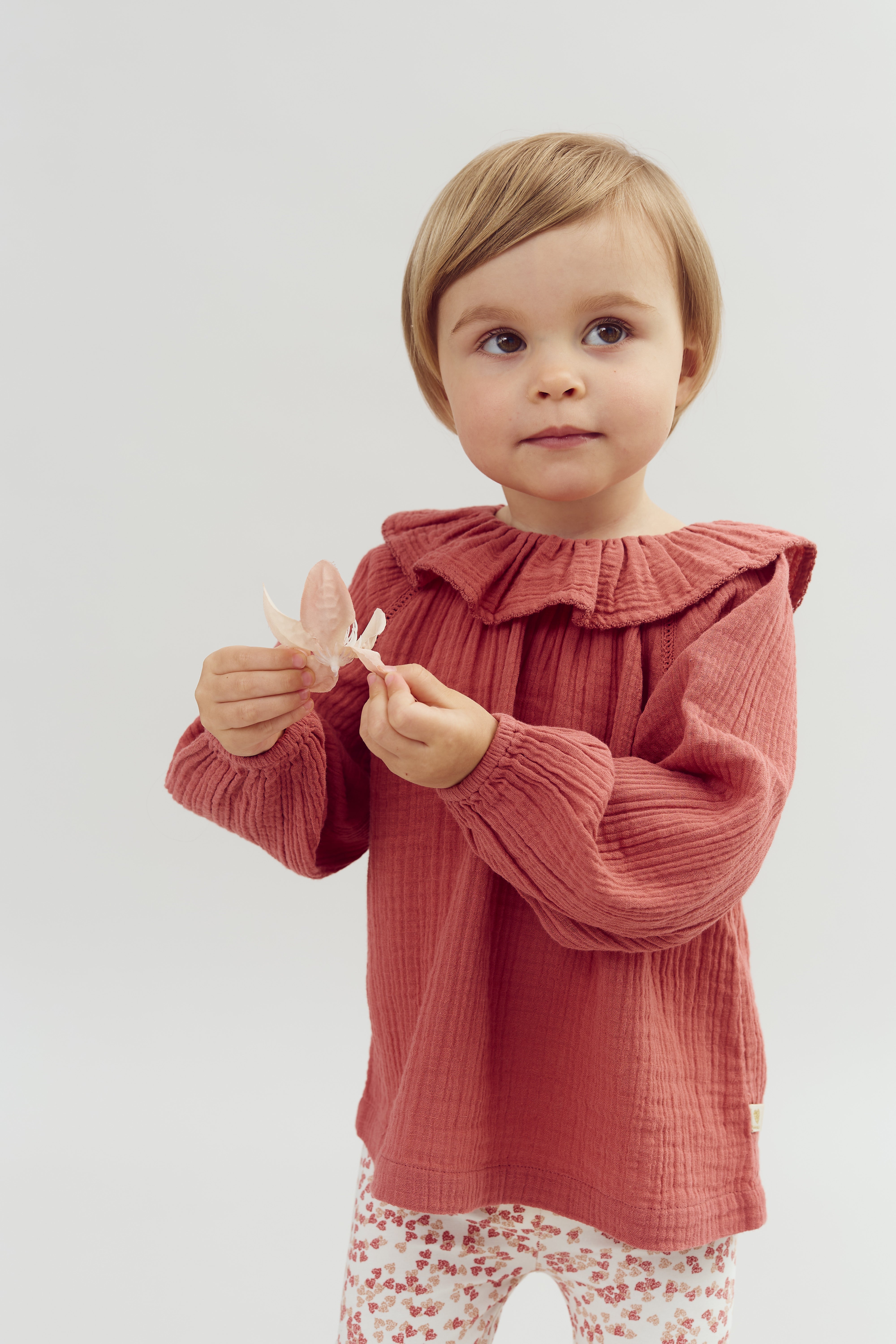 Child wearing a textured pink top, holding a delicate pink leaf, stands against a plain light background.