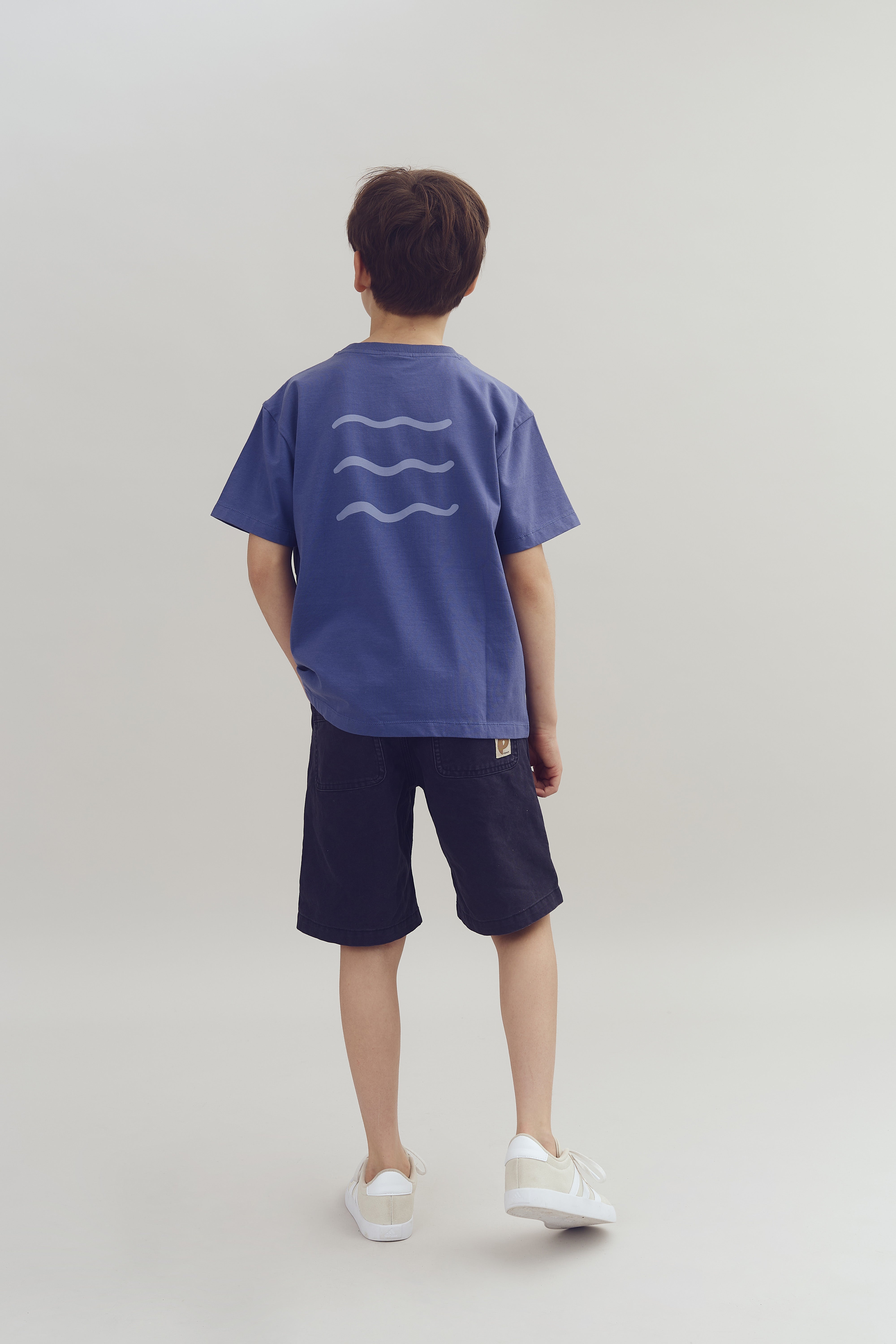 Ein Kind steht mit dem Rücken zur Kamera. Es trägt ein blaues T-Shirt mit drei Wellenlinien auf dem Rücken, dunkle Shorts und weiße Turnschuhe in einem neutralen Raum.