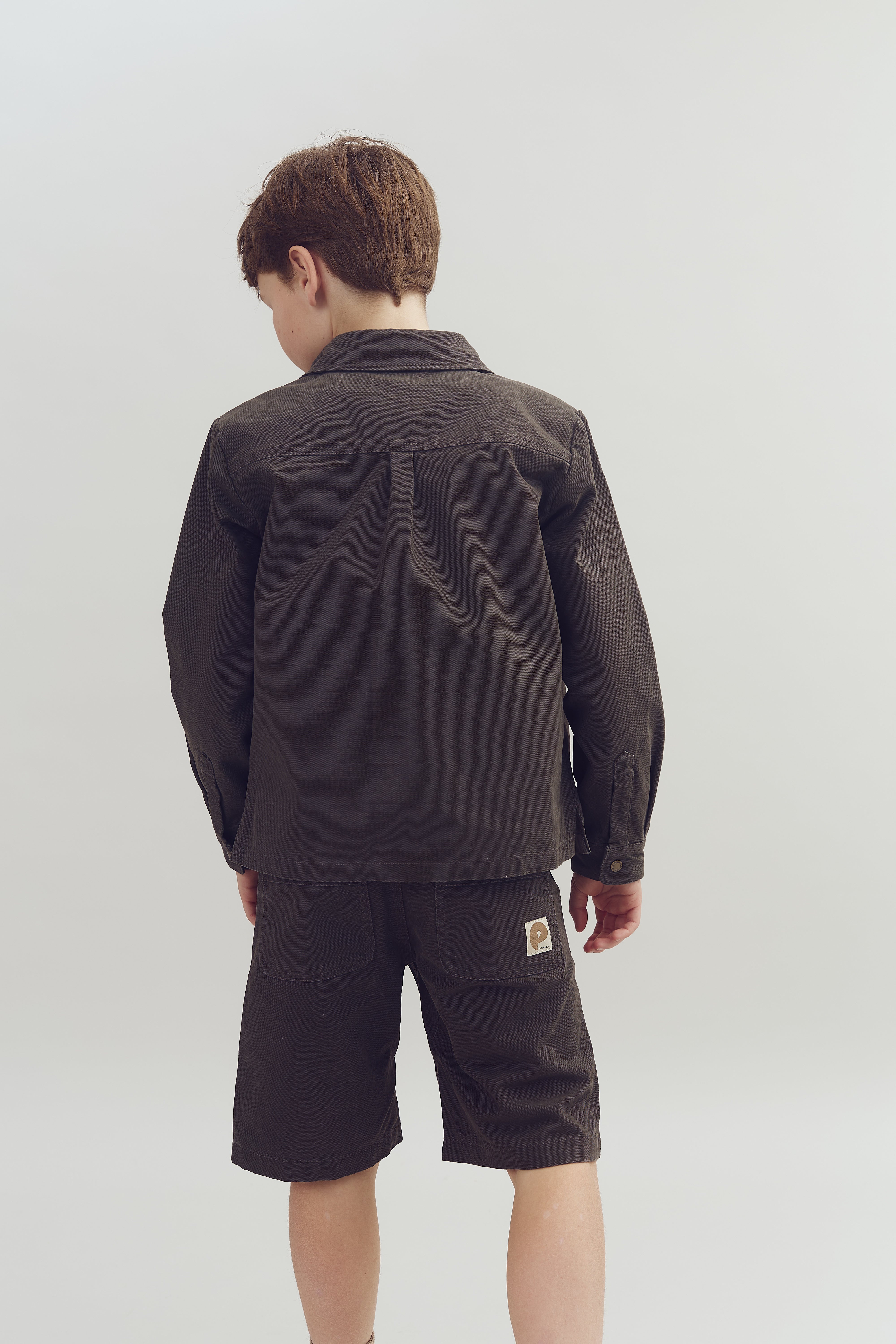 Ein Junge steht mit dem Rücken zur Kamera, trägt eine braune Jacke und Shorts in einem hellen Raum. Auf der Tasche der Shorts befindet sich ein eingenähtes Label.