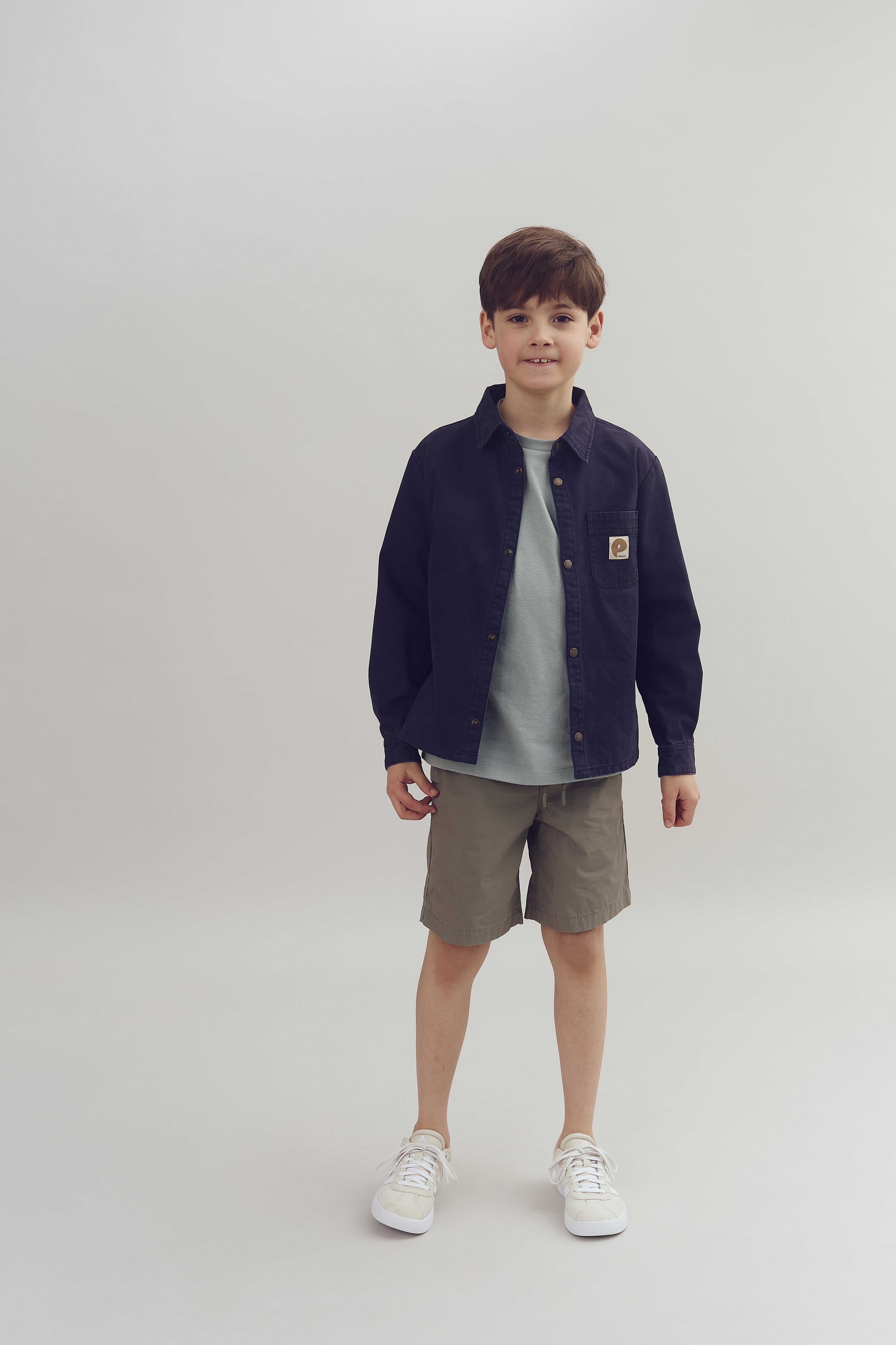 Ein Junge steht still, trägt eine dunkle Jacke, ein graues T-Shirt, beige Shorts und weiße Turnschuhe. Der Hintergrund ist einfarbig und hell.