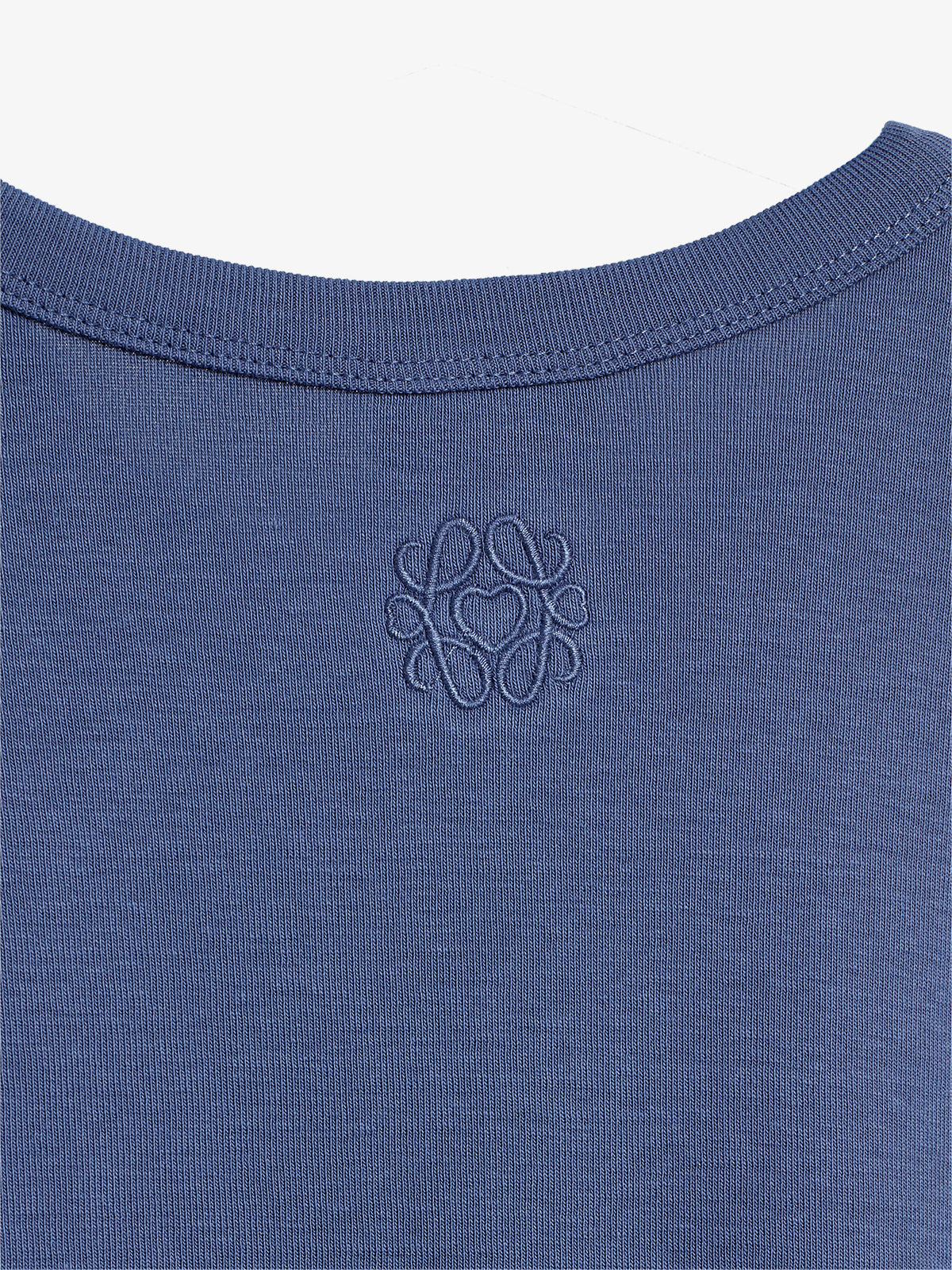 Ein blaues T-Shirt zeigt ein gesticktes Logo mit verschlungenem Muster im Nackenbereich. Die Struktur des Stoffs ist deutlich sichtbar und das Design wirkt minimalistisch.