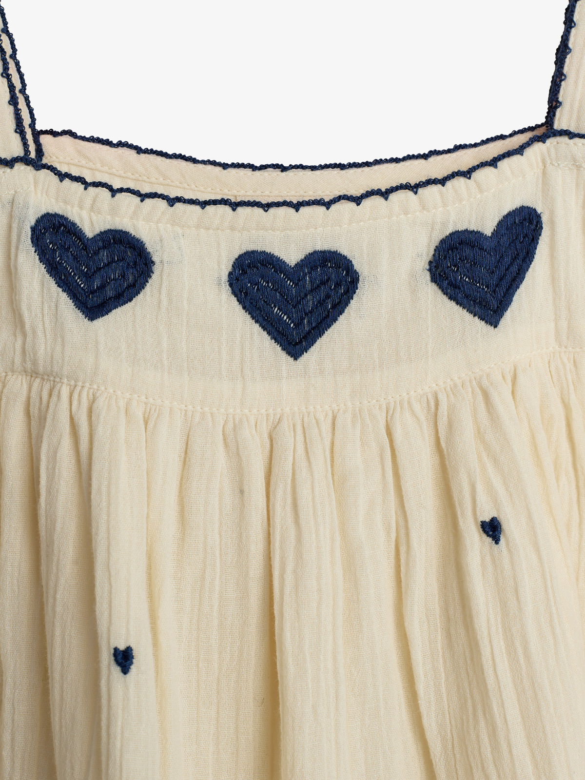 Ein beiges Kleid zeigt blaue gestickte Herzen in zwei Reihen. Die obere Reihe hat drei große Herzen, die untere kleinere Herzen. Es besteht aus leichtem Stoff.