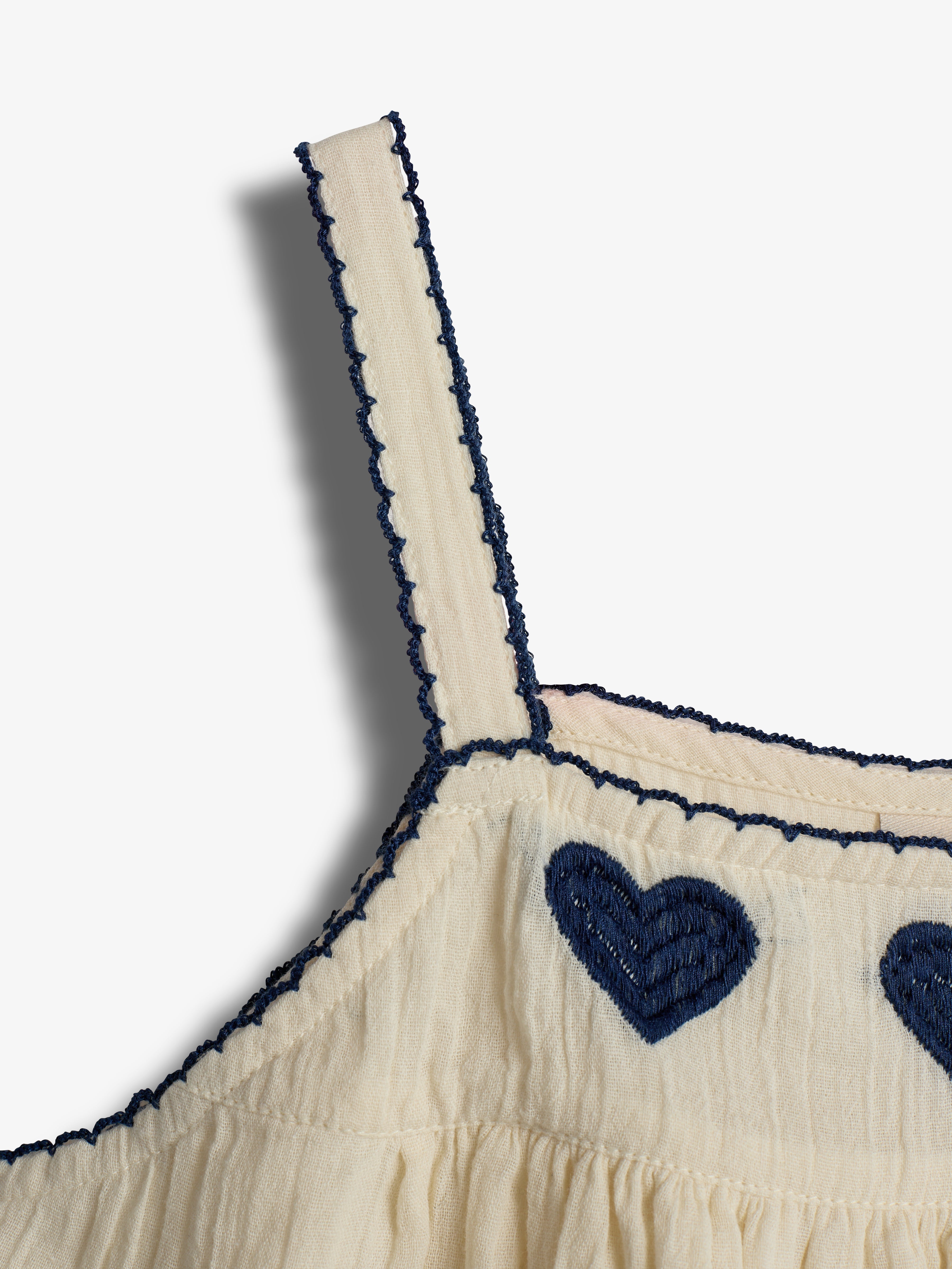 Cremefarbenes Kleid mit Spaghettiträgern. Dunkelblaue Herzstickereien zieren den oberen Teil. Der Saum ist mit dunkelblauen Fäden gesäumt. Schlichter Hintergrund in Weiß.