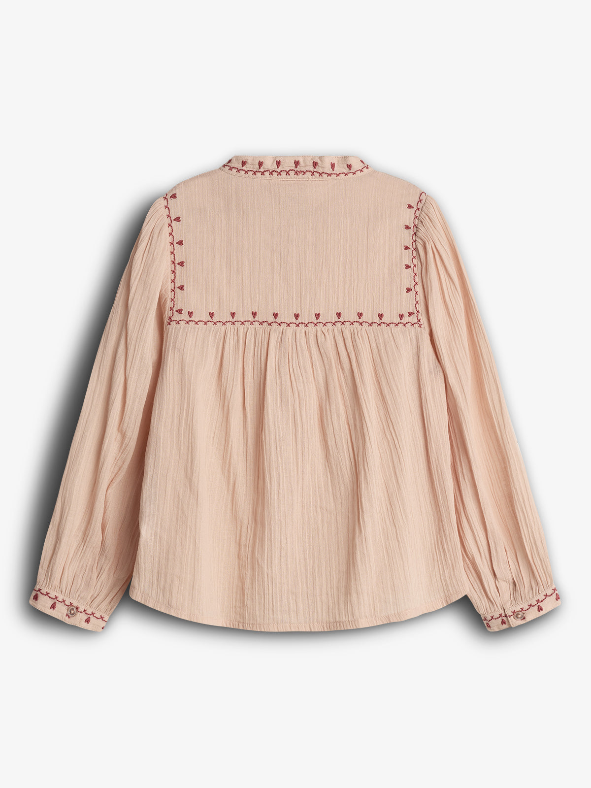 Eine beige Bluse mit roten Stickereien am Kragen und den Ärmeln. Die Bluse liegt auf einem weißen Hintergrund und zeigt eine geraffte Textur im Rückenteil.