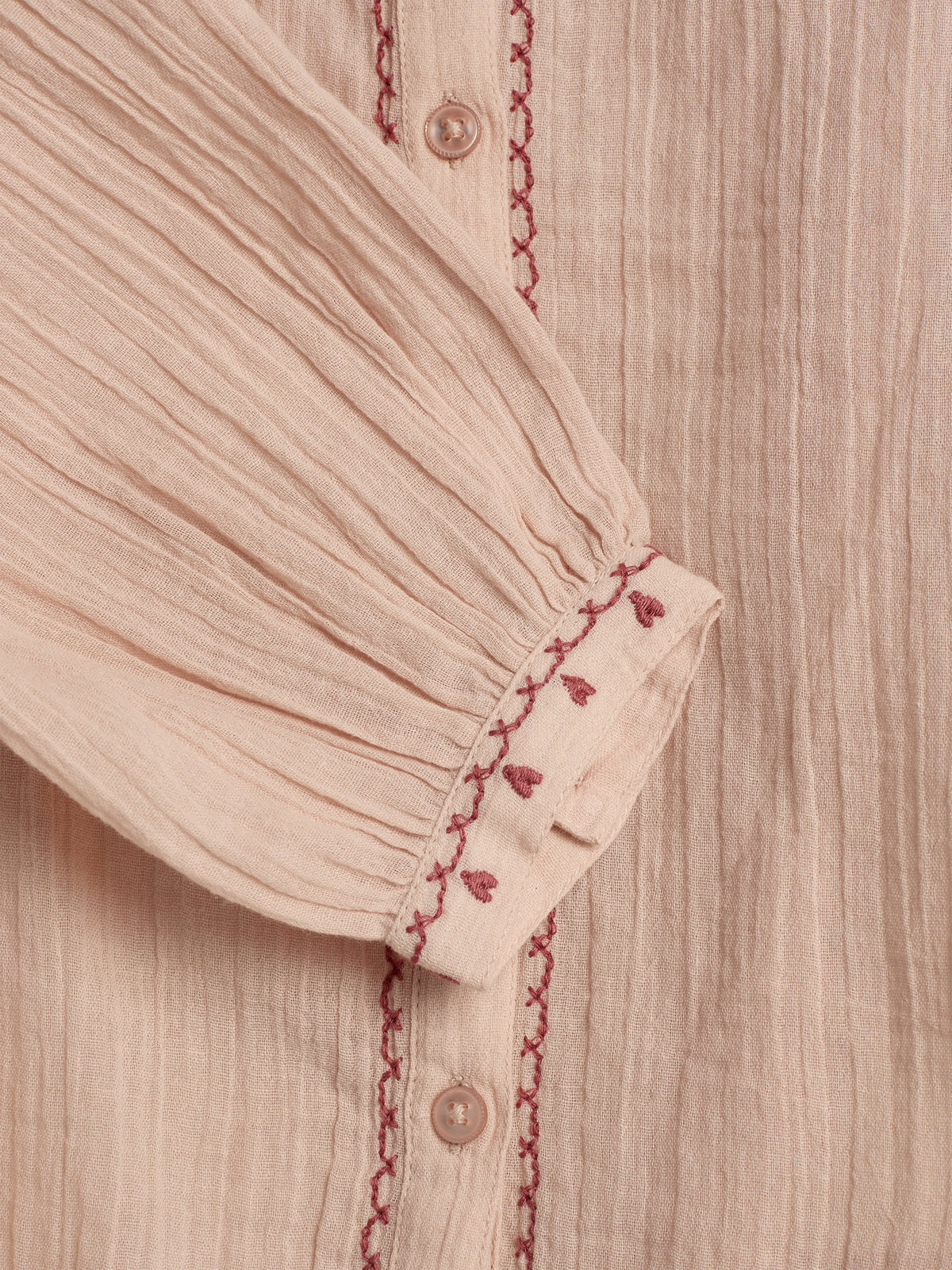 Beigefarbene Bluse mit rosa Stickerei und Knöpfen, die eine feine Textur aufweist. Der Ärmel ist gefaltet und zeigt delikate Muster, die einen handgefertigten Eindruck vermitteln.