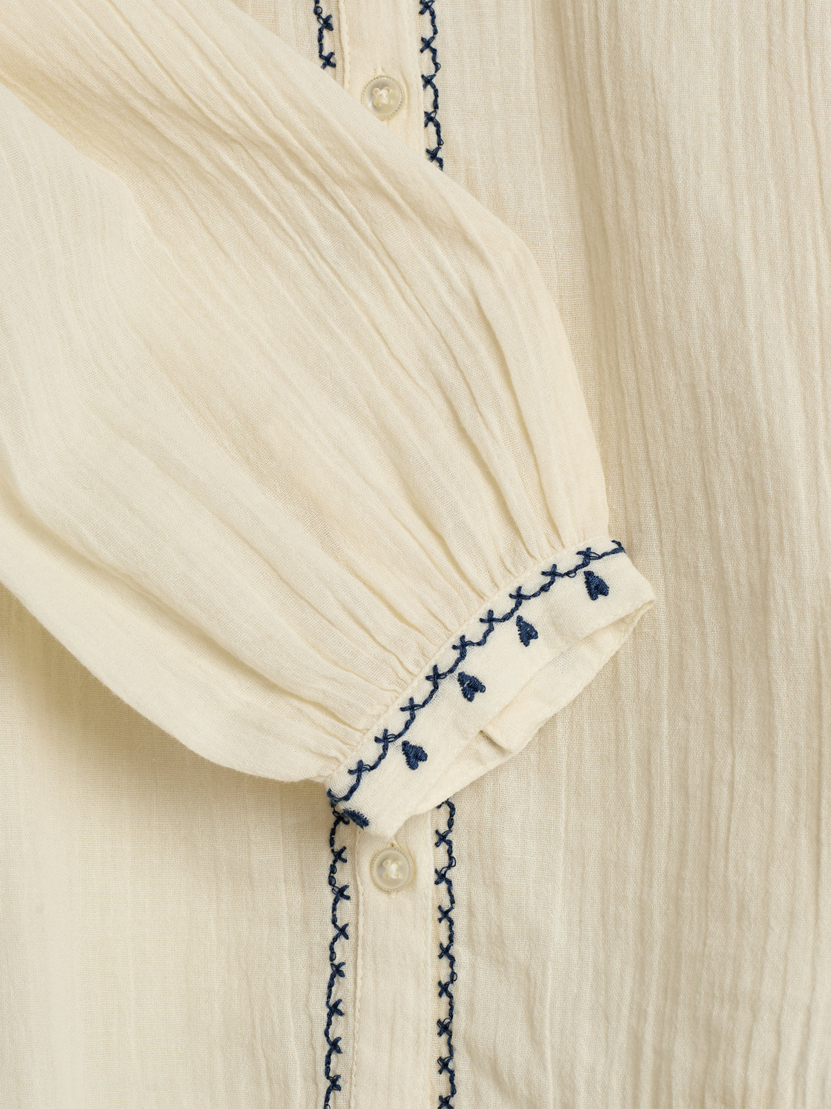 Eine beige Bluse mit blauen Kreuzstickereien ziert die Vorderseite und Manschette. In der Mitte befinden sich weiße Knöpfe. Der Stoff ist leicht strukturiert.