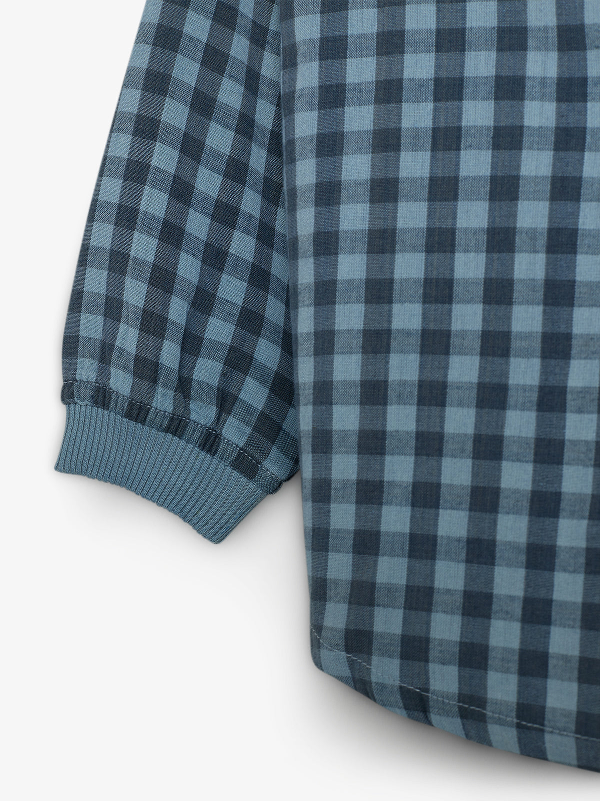 Ein Hemd mit blau-schwarzem Karomuster, dessen Ärmel mit einem elastischen Bündchen endet, liegt flach in einem neutralen, weißen Hintergrund.