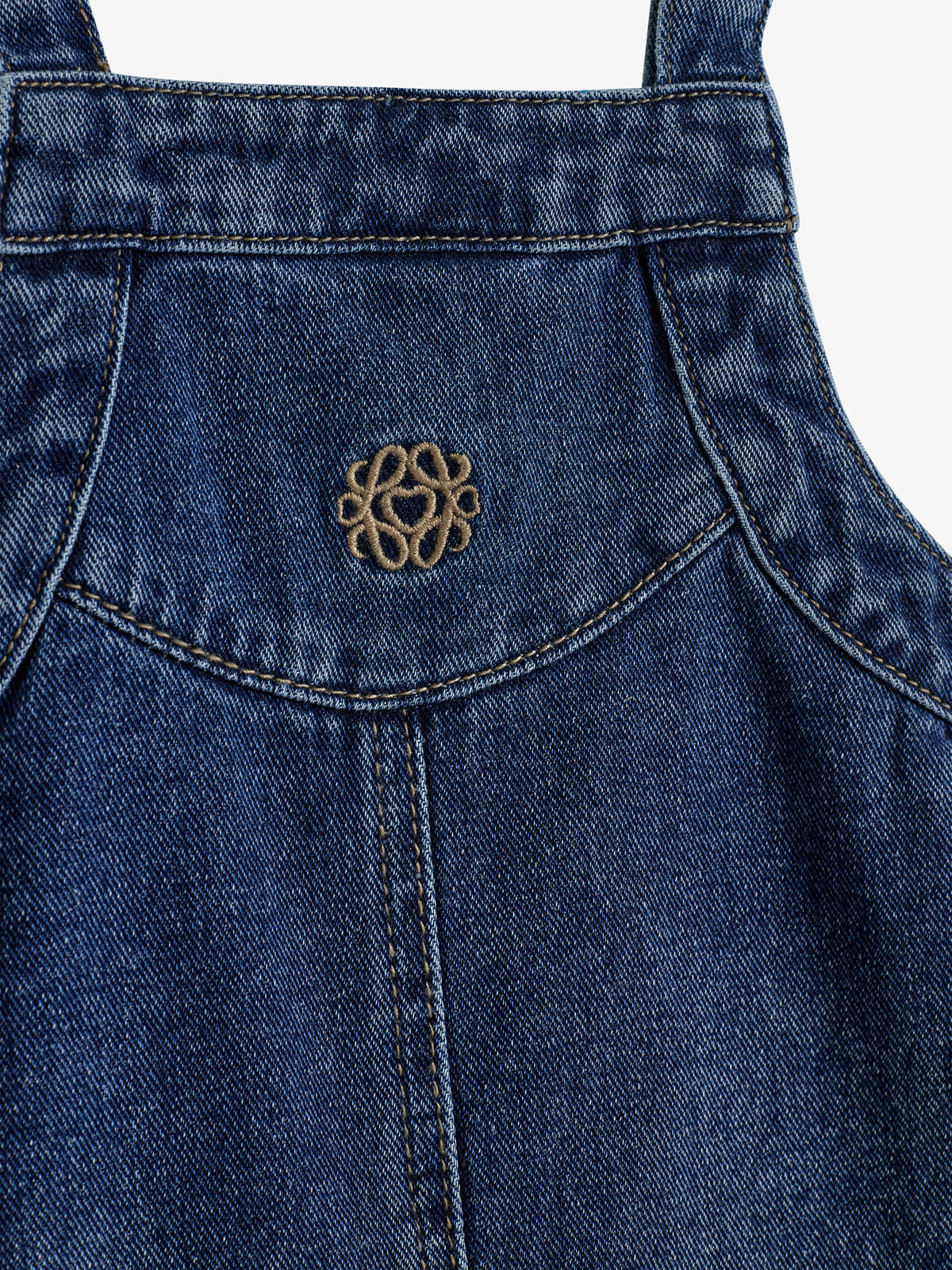 Jeansstoff mit Ziernaht und kleinem, stickähnlichem Logo in der Mitte, zeigt Detailansicht einer Latzhose vor einem einfachen Hintergrund.