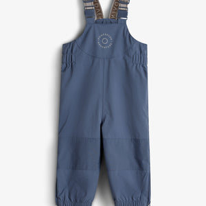 Die blaue Kinderlatzhose mit verstellbaren Trägern und elastischen Bündchen hat auf der Brust den Aufdruck "ROMPDELUX ADVENTURE". Sie hängt vor einem neutralen, weißen Hintergrund.