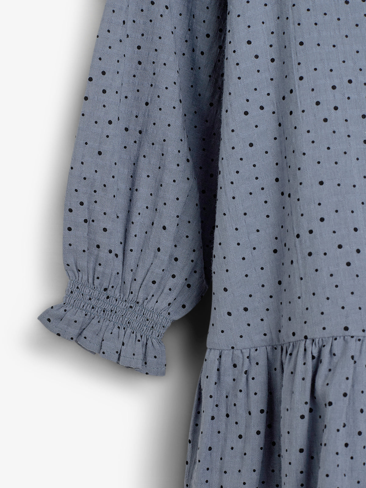 Graues Kleid mit schwarzen Punkten, zeigt bauschige Ärmel, die gerafft und elastisch sind. Der Stoff ist leicht und locker fallend, vor einem hellen Hintergrund.