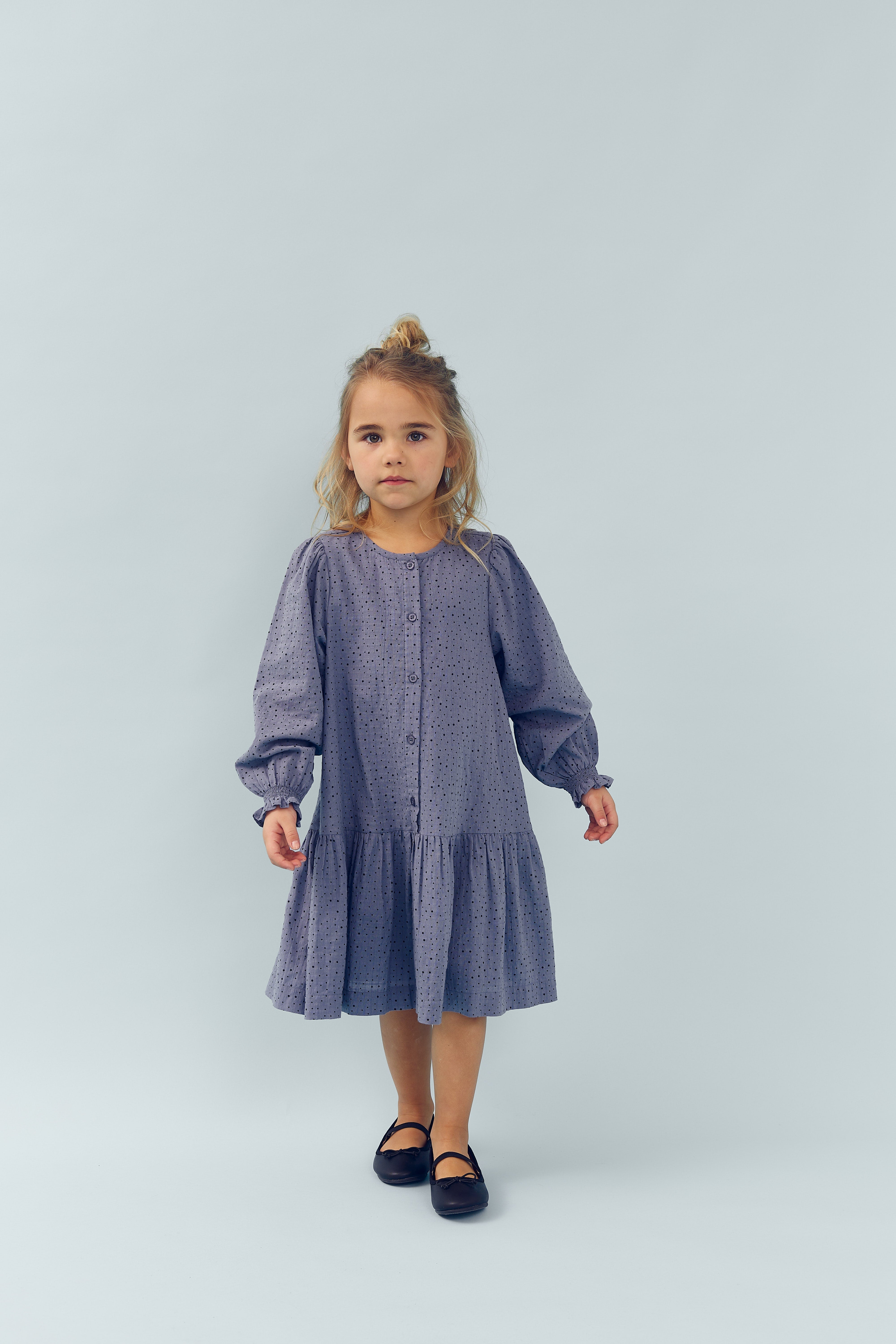 Ein kleines Mädchen steht ruhig und blickt geradeaus. Sie trägt ein langärmeliges, blaues Kleid und schwarze Schuhe, in einem hellen, neutralen Raum.