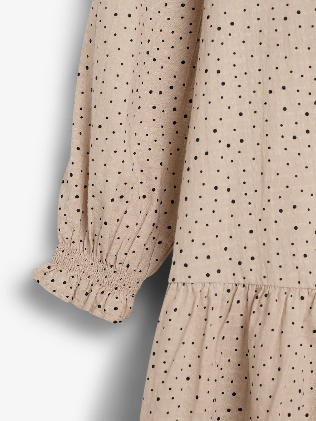 Ein beige Kleid mit schwarzen Punkten; der Ärmel ist gesmokt und leicht gerafft. Der Hintergrund ist schlicht und einfarbig, was den Fokus auf das Muster lenkt.