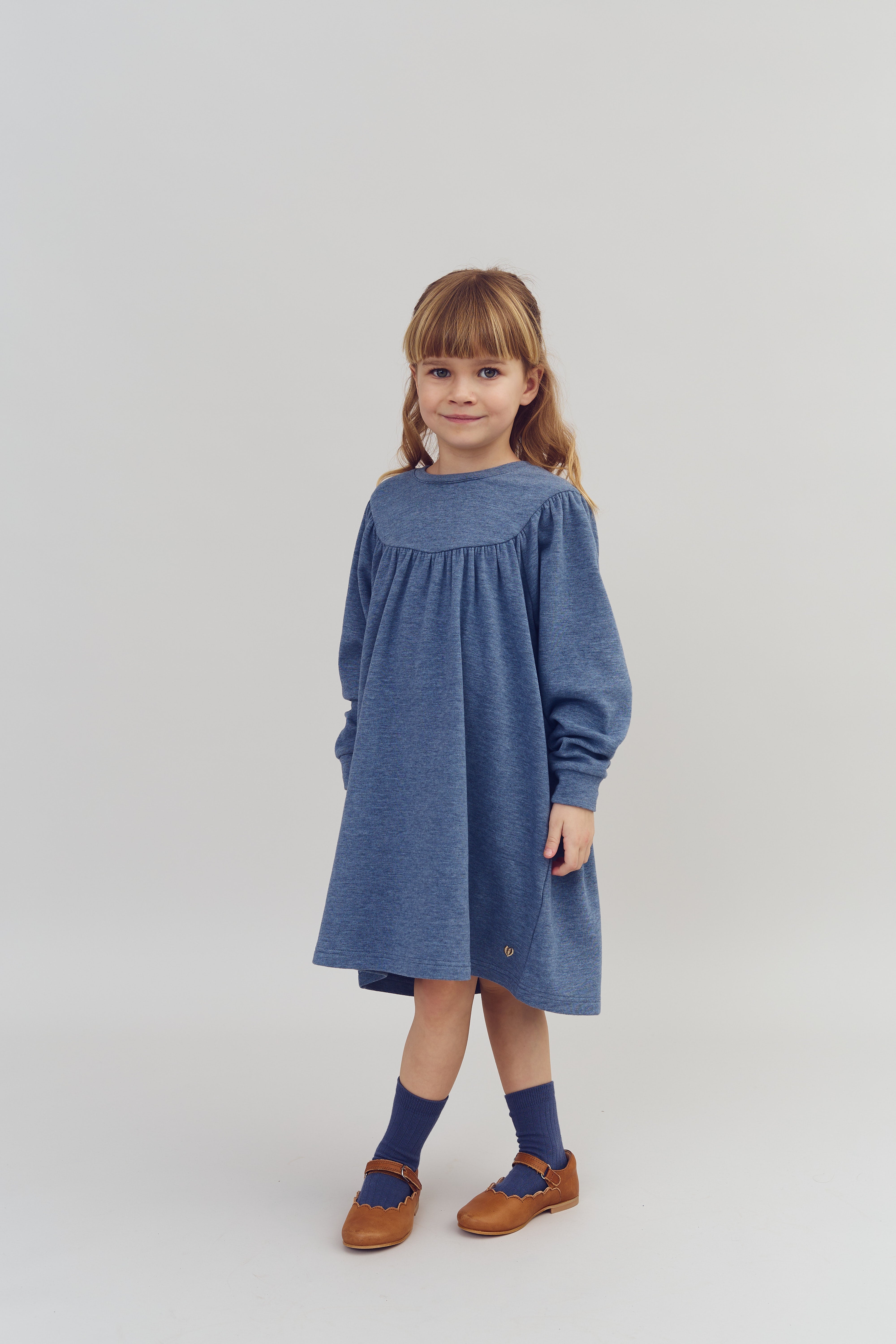 Mädchen steht in einem blauen Kleid mit langen Ärmeln, braunen Schuhen und blauen Socken vor neutralem Hintergrund, lächelt dezent.