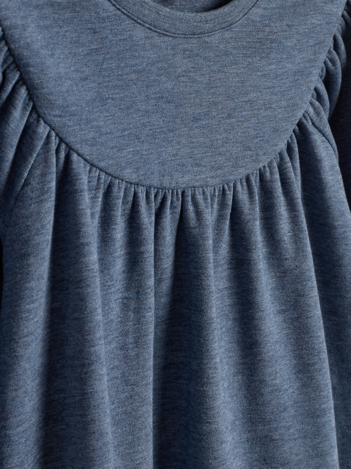 Ein blaues Kleidungsstück aus weichem Stoff mit gerafften Details bildet Falten. Es hängt in einem ruhigen, neutralen Umfeld und zeigt die Textur und den fließenden Stil des Stoffes.