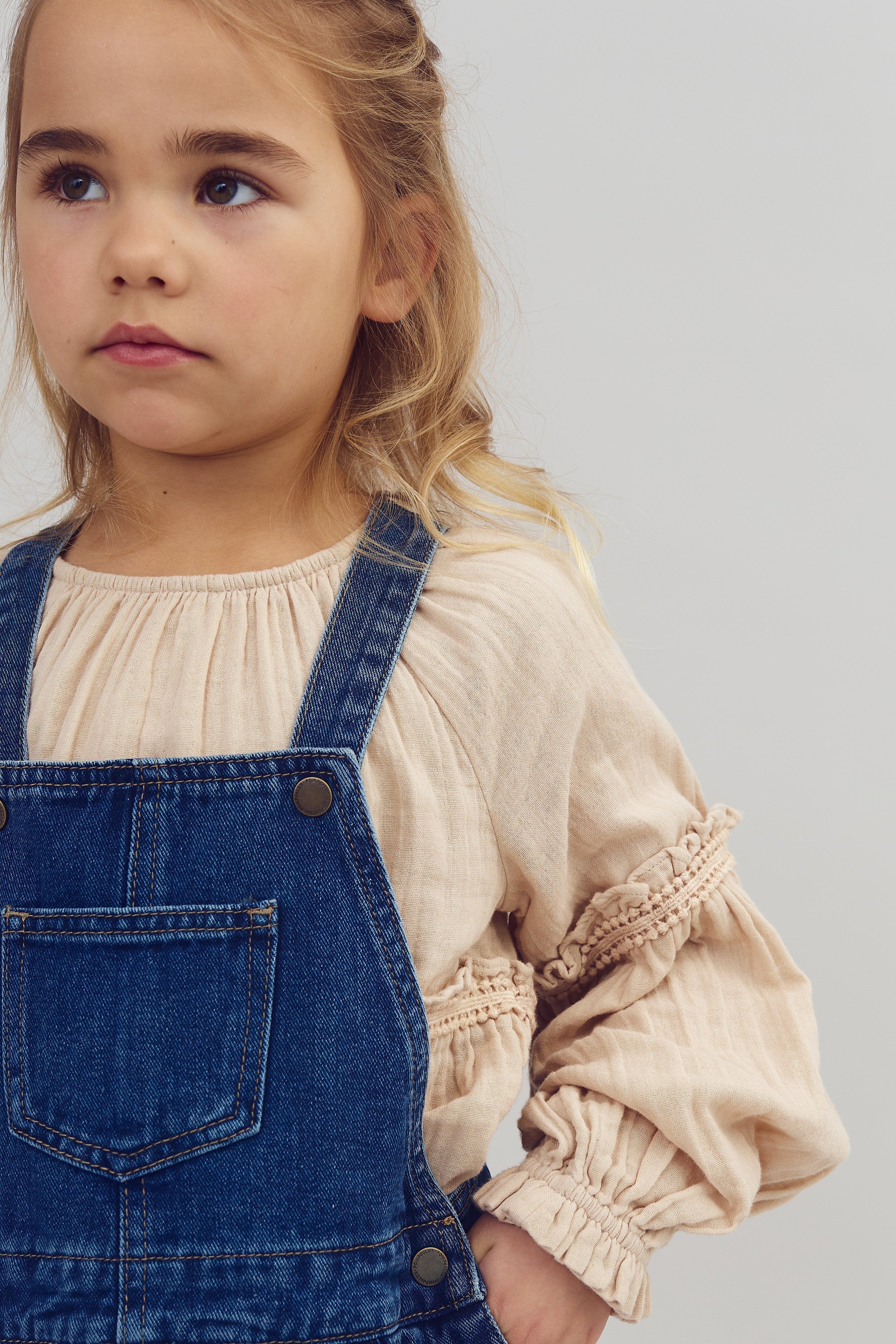 Ein Kind trägt eine Jeanslatzhose über einem beigen, langärmeligen Oberteil. Das Kind blickt nach links und trägt sein Haar locker. Der Hintergrund ist neutral.