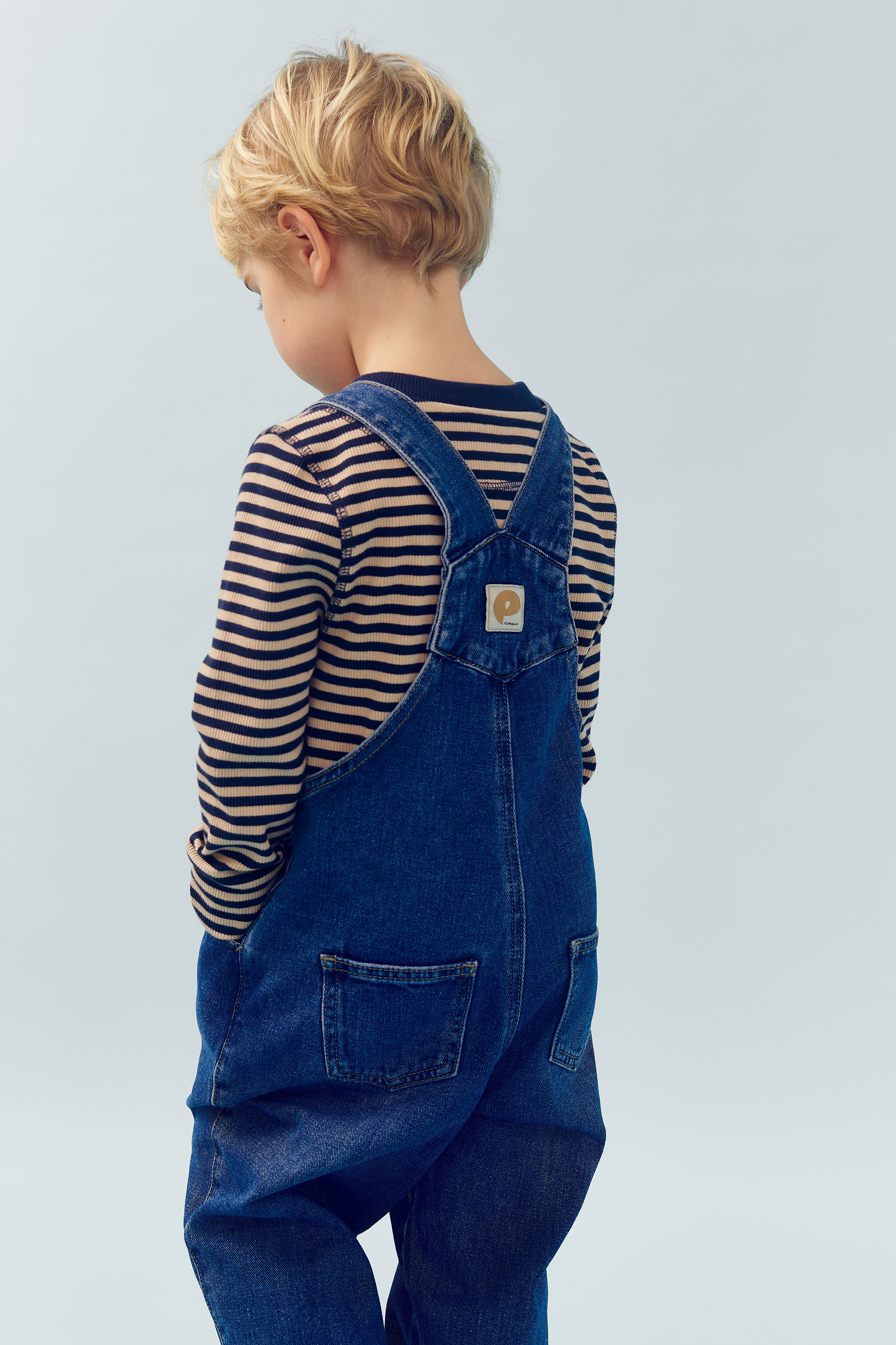 Ein Kind mit blonden Haaren trägt eine gestreifte Haube und blaue Jeans-Latzhose mit aufgenähtem Etikett. Es steht seitlich in einem hellblauen Raum.